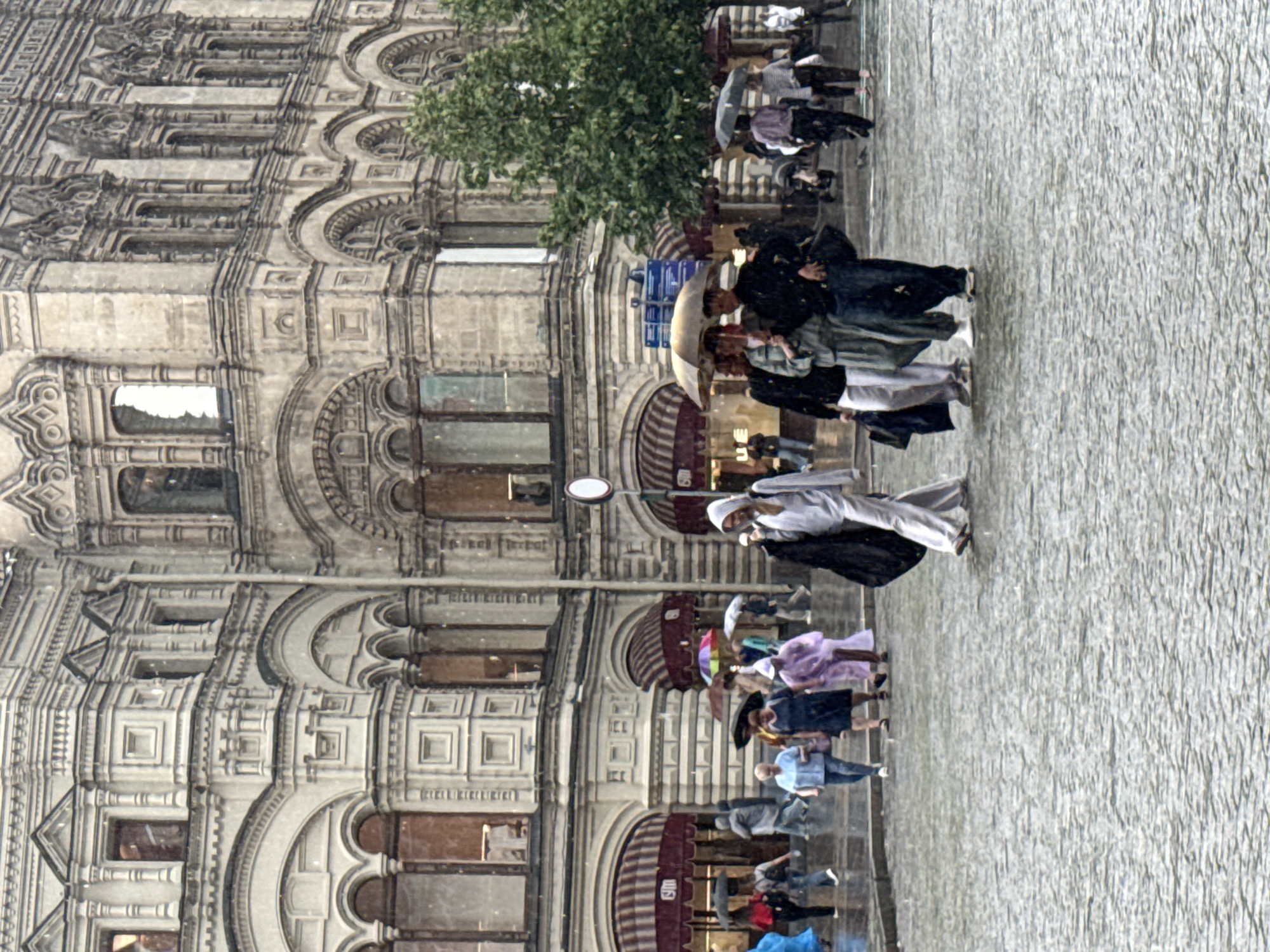 This image depicts a rainy day scene in Red Square, Moscow, Russia. The grand facade of the GUM department store serves as the backdrop, its ornate architectural details visible despite the inclement weather. Several people are present, walking on the wet cobblestone pavement, many holding umbrellas of various colors. Some are dressed in raincoats and casual clothing. The rain is actively falling, creating a misty effect and making the ground appear slick. The atmosphere is that of a typical bustling urban environment enduring a downpour.