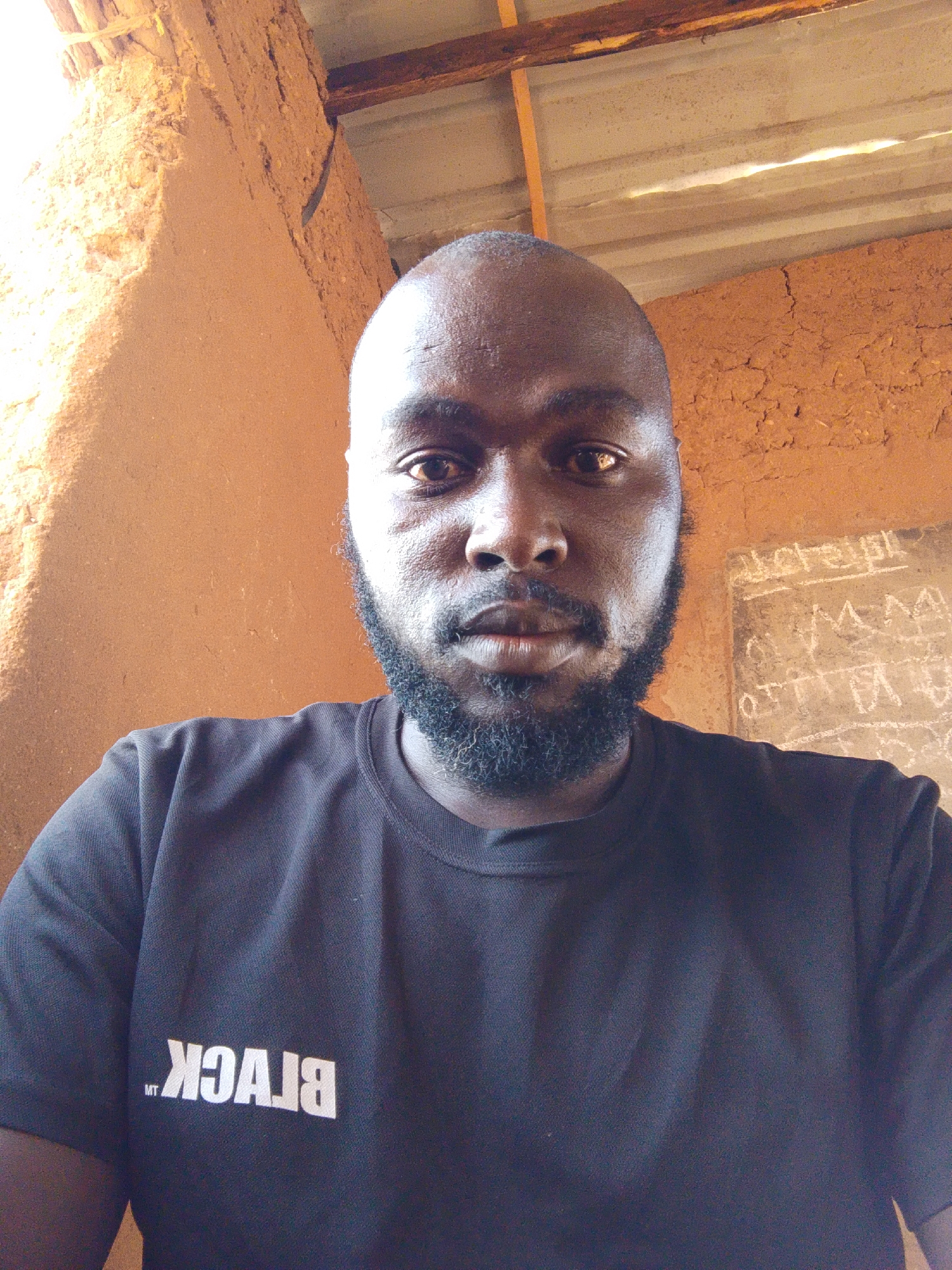 A close-up, self-portrait of a dark-skinned man with a shaved head and a full, short, dark beard, wearing a black t-shirt with the word "BLACK" printed in white and reversed. The background consists of a rough, reddish-brown wall and a partially visible ceiling made of corrugated metal sheets and wooden beams. There is also a wall with some written characters. The location is presumably Mulke Barikin Sayejo, Nigeria. The man is looking directly at the camera.