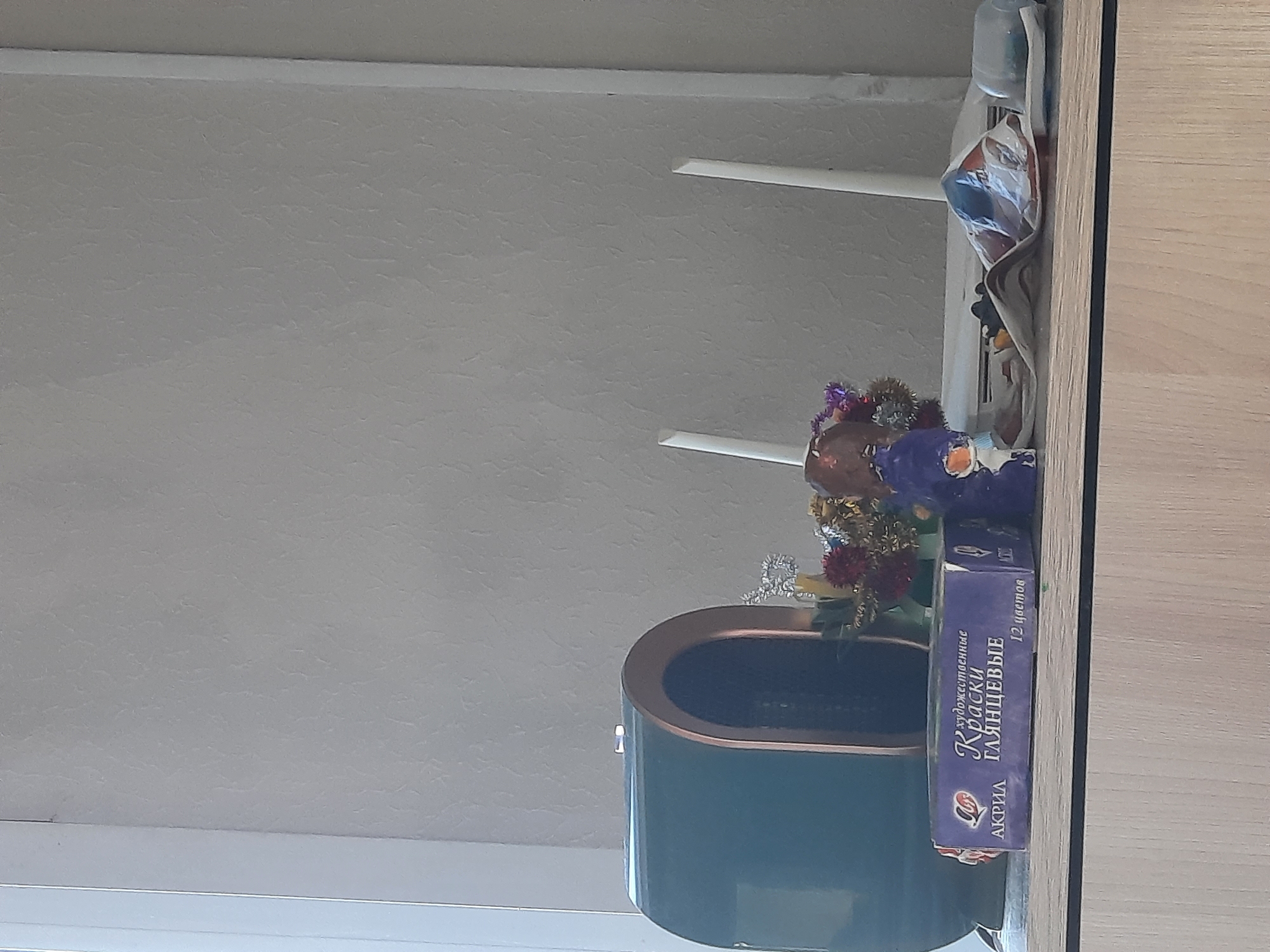 The image shows a view of a room, likely in a residential setting. A teal-colored, oval-shaped object, possibly a container or vase, sits near a window. Inside the container, there are floral decorations. Next to the container is a box labeled "КРАСКИ ГЛЯНЦЕВЫЕ," which translates to "GLOSSY PAINTS" in English. The box indicates that the paints are artistic and come in 12 colors. The text "АКРИЛ" is also present, indicating that the paints are acrylic-based. Above these objects, there are two white ceiling fan blades partially visible. A plastic bottle is visible near the top of the frame. The photograph is taken indoors, and the location is not clearly indicated in the city of Krasnogorskoye, Russia.