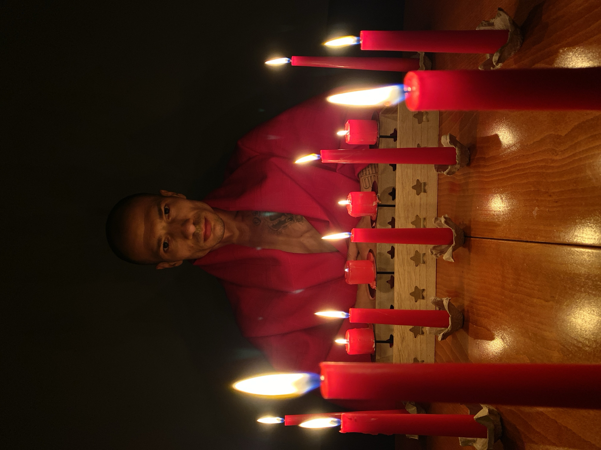 The original image, when viewed in its correct upright orientation, captures a man and a collection of lit red candles in a dimly lit indoor setting.
On the left side of the frame, a man with a shaved head or very short dark hair, light brown skin, and a faint mustache is visible. He is wearing a vibrant red robe or garment and looks directly at the camera with a subtle, pleasant smile. A tattoo is discernible on his left shoulder, partially covered by his clothing.
To his right, a prominent array of red candles of varying sizes, both slender and thicker, are burning brightly. These candles are arranged in a linear fashion within a wooden stand, which is adorned with decorative star-shaped cutouts. Each candle is held in place by what appears to be a small, crinkled piece of foil or paper acting as a holder. The candles rest on a polished wooden table, which vividly reflects the warm, flickering flames and glowing light. The scene is primarily illuminated by the intense light emitted from the candles, suggesting a nighttime setting or a very dark room. The background behind the man is uniformly deep black, creating a stark contrast with the brightly lit foreground.
The arrangement of numerous lit candles hints at a ceremonial, festive, or spiritual event. No visible text is present in the image.