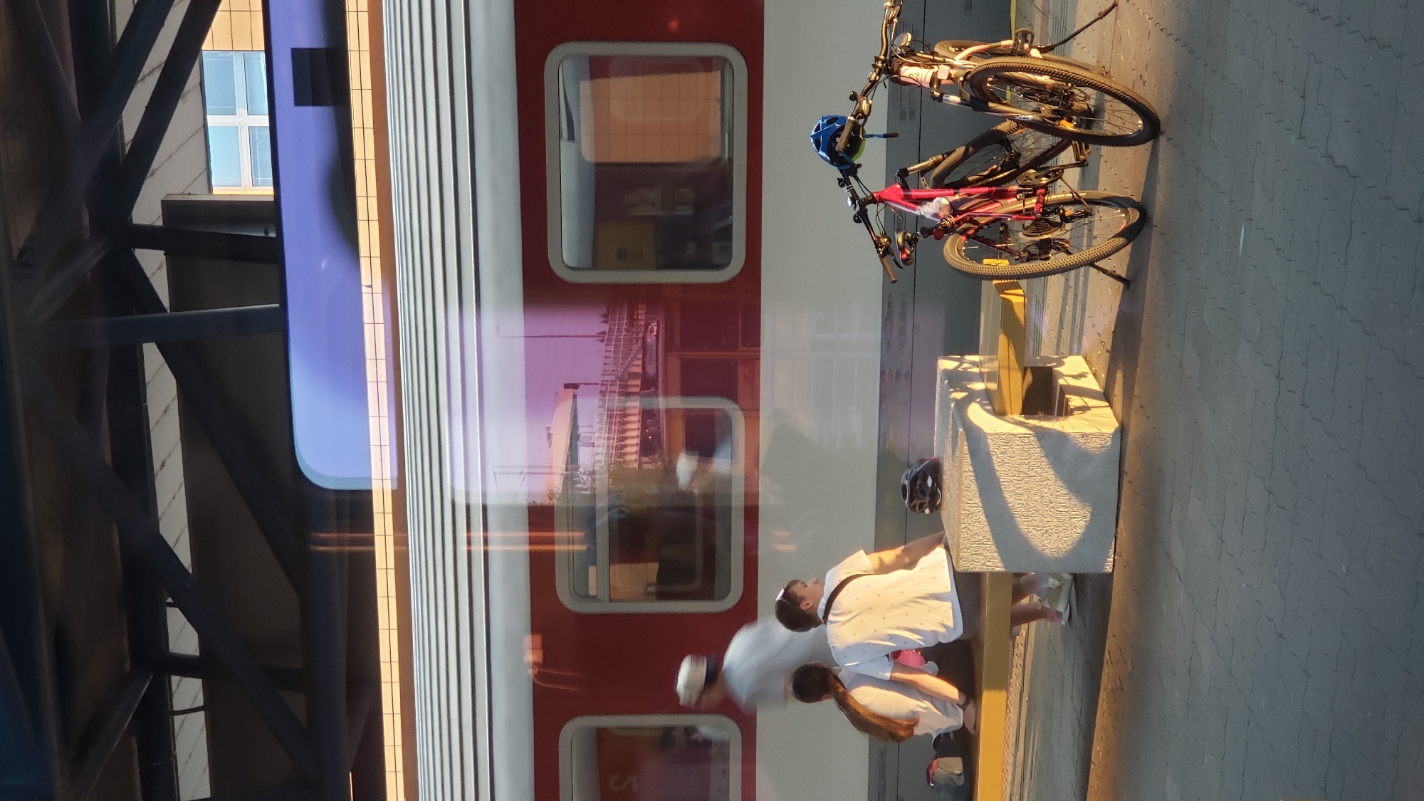 This image depicts a scene at the train station in Poprad, Slovakia, during what appears to be late afternoon or early evening, given the long shadows and warm lighting.
In the foreground, two bicycles are parked. One is a pink mountain bike with a blue helmet attached to its handlebars. The other is a black bicycle, partially obscured, also with a helmet on its handlebars.
To the left of the bicycles, a modern concrete bench with a wooden seat is situated. On this bench, three people are seated. Two individuals, a male and a female, are sitting close together, facing the train. The female has long dark hair and is wearing a white t-shirt. The male is wearing a white t-shirt with a subtle pattern. A third person, seemingly a man, is standing behind them, partially obscured by the seated individuals, wearing a white shirt and a light-colored cap, and appears to be moving.
In the background, a red and white train is stopped at the platform. The train's windows reflect the surrounding environment, including overhead structures and what appears to be an elevated pedestrian walkway or bridge. The train has a distinctive blue stripe along its upper portion. The platform itself is paved with grey interlocking tiles.
Overhead, a modern train station roof structure is visible, supported by dark metal beams. In the far background, a building with a grid-like window pattern is present. The overall impression is one of a tranquil moment at a public transportation hub.