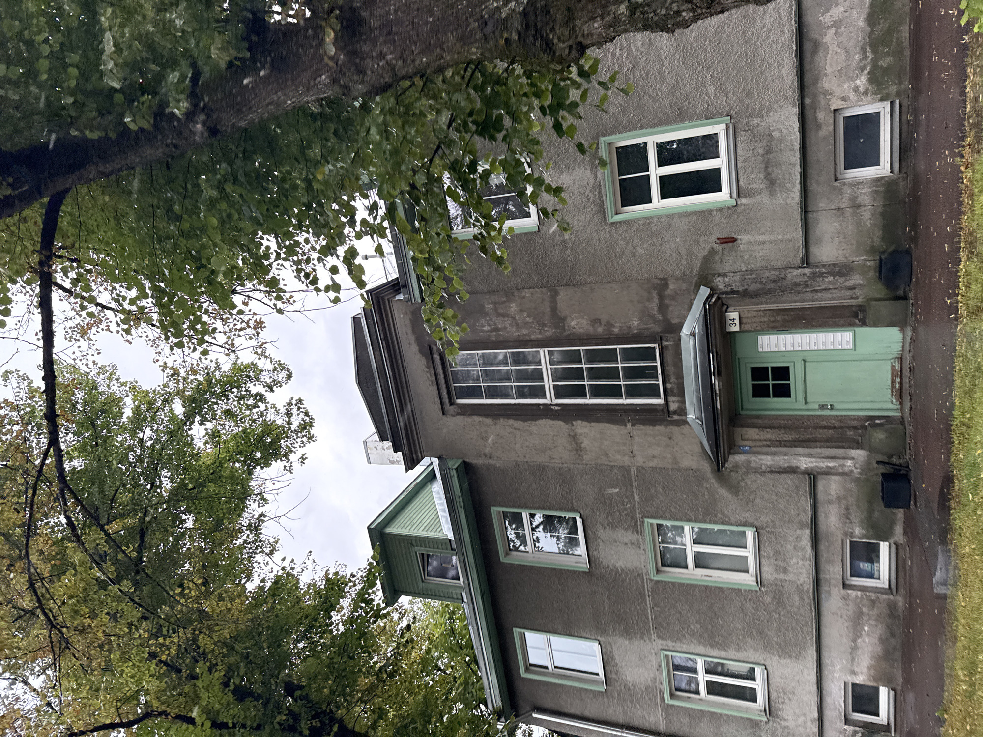 The image shows the facade of a grey, multi-story building with a green, pitched roof dormer on the top left. The building has multiple windows, most of which are white-framed and rectangular, though one on the upper right is taller and multi-paned. The building's entrance features a bright green door with a small window in its upper half, and a sign with the number "34" above it. Two dark bins are placed near the entrance. The building is partially obscured by the branches of two large trees with green and yellowing leaves, suggesting an autumn setting. The ground in front of the building is covered with a mixture of asphalt and grass, with fallen leaves scattered on the ground. The sky is overcast. The architectural style suggests the building is located in the Kalamaja district of Tallinn, Estonia, known for its historic wooden houses and pre-war apartment buildings.