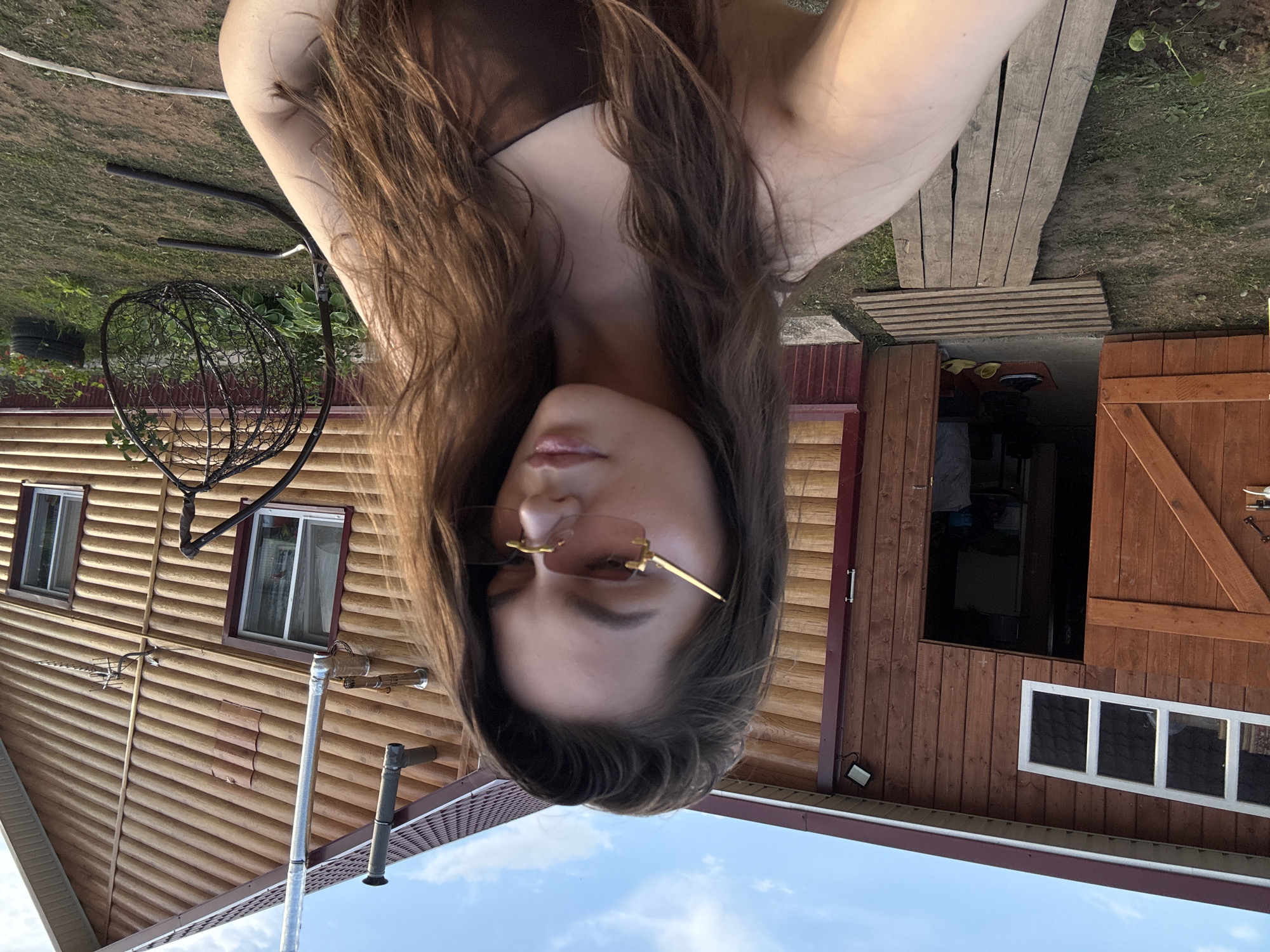 A young Caucasian woman with long, brown hair and light skin, wearing rimless sunglasses with gold frames and a dark brown top, is captured in a selfie. She is positioned in front of a rustic, wooden building with a red-brown roof. The building features a wide, open doorway with a wooden door ajar, revealing the interior of what appears to be a shed or garage. Several windows are visible on the building's facade, one of which has a light fixture above it. A wooden walkway leads to the entrance. To the right of the building, a black, hanging egg chair is visible on a metal stand, amidst green grass and foliage. The sky above is partly cloudy. The scene appears to be set in a rural or semi-rural location, possibly a dacha or country house, given the rustic architecture and outdoor setting.