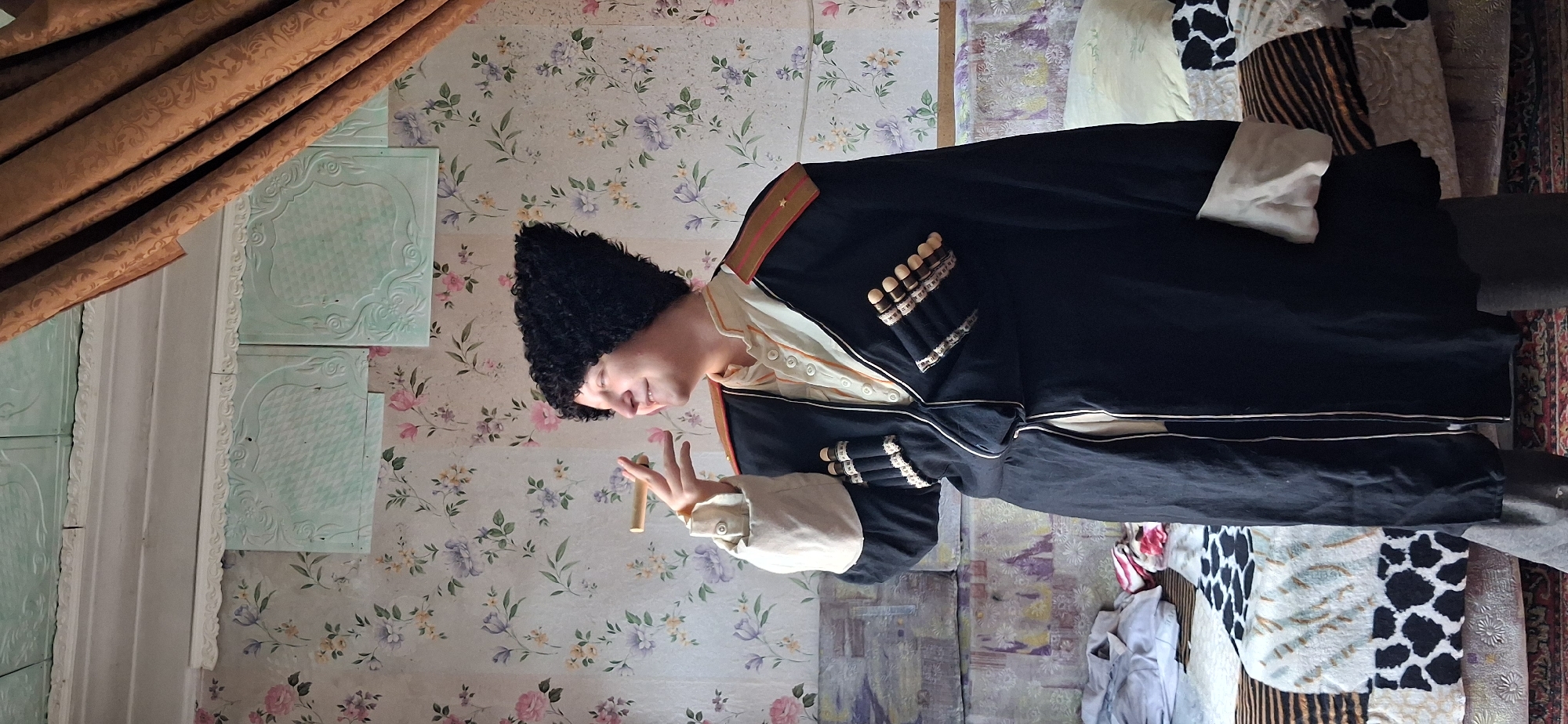 In the city of Petropavl City Administration, Kazakhstan, a person is depicted indoors, standing in front of a floral-patterned wallpaper. The person is wearing a traditional black fur hat and a dark coat adorned with epaulets and what appear to be decorative cartridges. The coat has light-colored trim along its opening and cuffs. The person is holding a cigar to their lips and smiling. In the background, a bed with patterned bedding is visible, along with a curtained window. The room's ceiling has decorative panels.