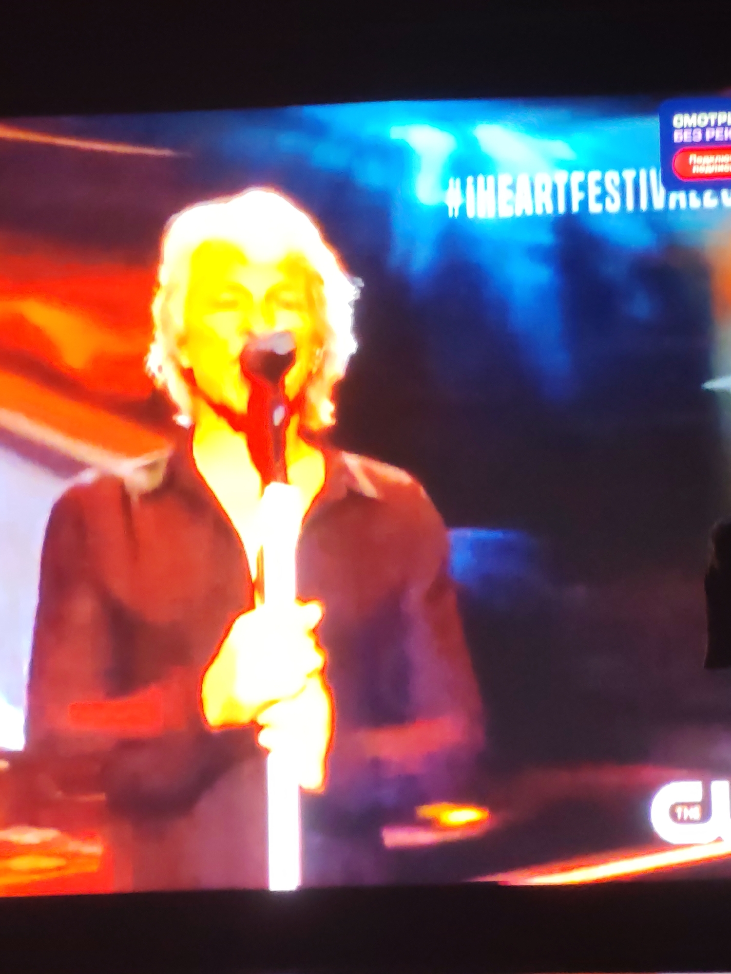 The image is a screenshot of a broadcast, likely from television, showing a performer singing into a microphone. The performer has light-colored, possibly blonde, hair and is wearing a dark-colored shirt. They are on a stage with dramatic lighting, featuring reds, oranges, and blues. Text is visible on the screen, including "#IHEARTFESTIVAL" and what appears to be Russian text related to viewing or subscribing. In the lower right corner, a logo that resembles "THE CW" is partially visible. The image quality is poor, with a significant amount of blur and pixelation, making it difficult to discern fine details. There is no information or visual cues within the image that would indicate the location in the city of Vladimir, Russia.
