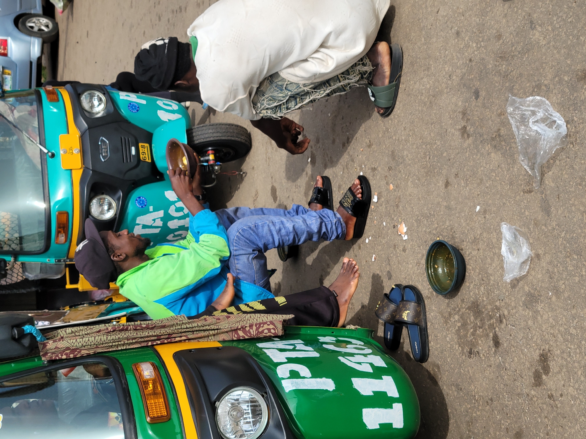 In Gura Top, Nigeria, two men are seated outdoors near a bright green and turquoise auto-rickshaw. One man, wearing a black baseball cap and a green and blue jacket, is holding a small bowl with food. The other man, wearing a white shirt and patterned pants, is crouched down, looking at the first man. Scattered on the ground around them are pieces of broken eggshells and a crumpled plastic bag. A pair of Louis Vuitton inspired sandals are placed near the auto-rickshaw. The ground is paved and appears to be a street or marketplace.