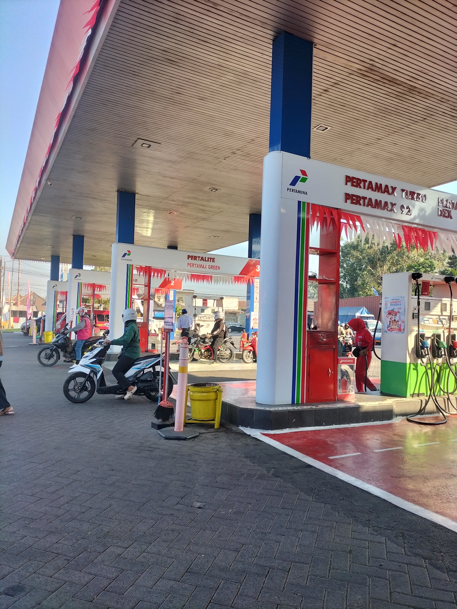 The image captures a scene at a Pertamina gas station in Mojosari, Indonesia. The view is from ground level, looking towards several gas pumps under a large canopy. The canopy has a wooden plank ceiling and is supported by blue pillars. Red and white pennant banners are strung along the edge of the canopy and also decorate the front of the gas pump structures.
In the foreground, the ground is paved with dark gray interlocking bricks. A bright yellow trash can with a pink pole stands on the brick paving, accompanied by a broom.
Several people on motorcycles are present. One person in a green jacket and white helmet is seated on a white scooter, facing away from the camera. Another person in a red jacket and helmet is on a black motorcycle to the left. Further back, another person on a motorcycle is visible near a gas pump.
The gas pump islands are white with colored vertical stripes (blue, green, red). Signage indicates different fuel types, including "PERTAMAX TURBO," "PERTAMAX $2," and "PERTALITE PERTAMAX GREEN." One of the pump structures has a red cabinet on its side. A person in a red uniform and headscarf is standing near this cabinet, holding a tool. Another gas pump to the right has green detailing and visible fuel hoses.
In the background, there are trees, buildings, and other vehicles, suggesting a typical urban or suburban environment. The sky is clear and blue. The lighting indicates it is daytime, likely morning or afternoon.