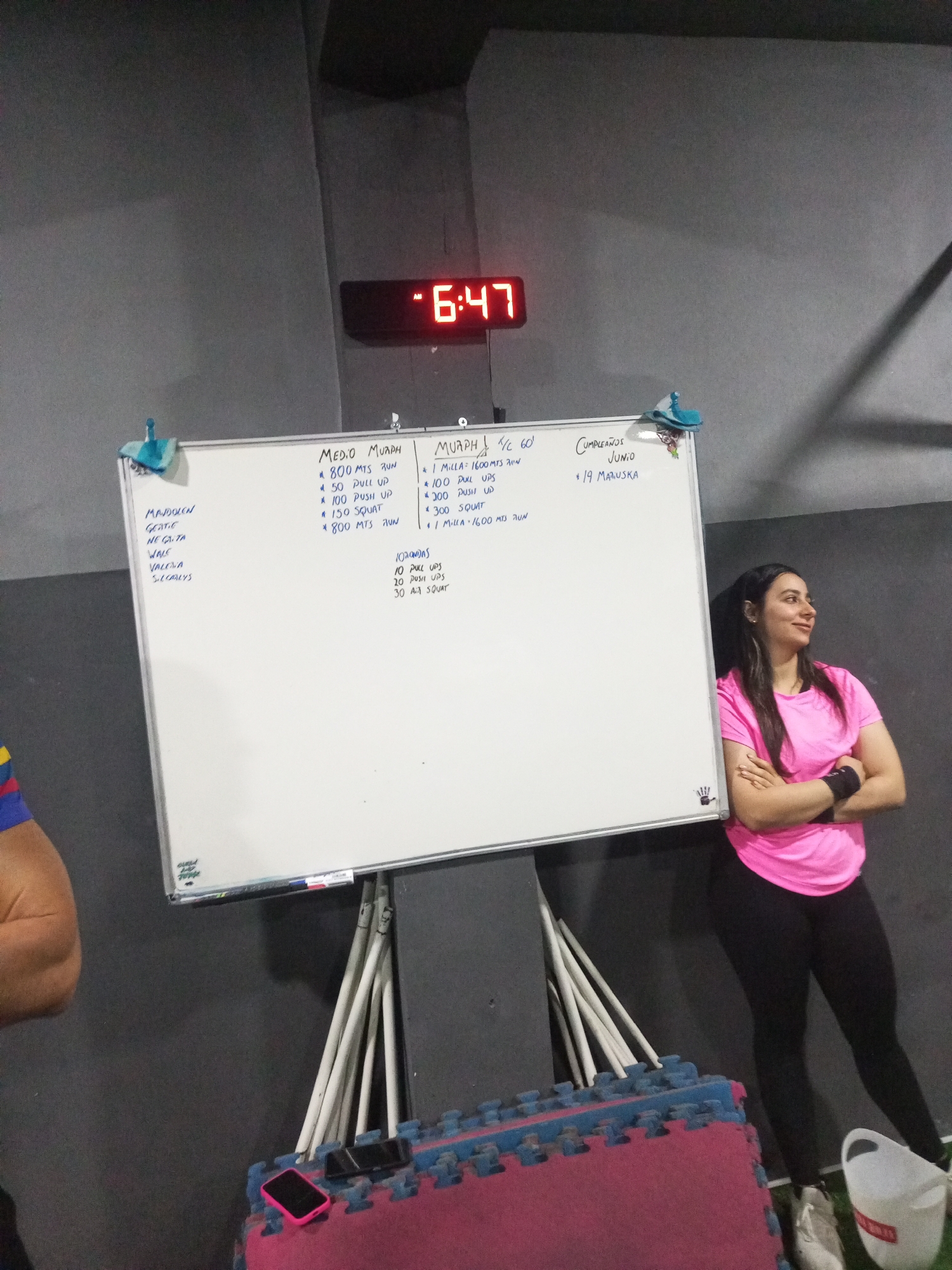 The image depicts a group of people in what appears to be a gym or fitness center. A whiteboard on the left side of the frame lists various exercises and workout routines. A digital clock on the wall displays the time "6:47". A woman in a pink shirt and black leggings is standing to the right of the whiteboard, leaning against it with her arms crossed. Several mats and gym equipment are visible in the foreground. The environment appears to be indoors, with a dark gray wall and a black ceiling. The overall lighting suggests it might be evening or a dimly lit indoor space. The location is identified as Juan Griego, Venezuela.