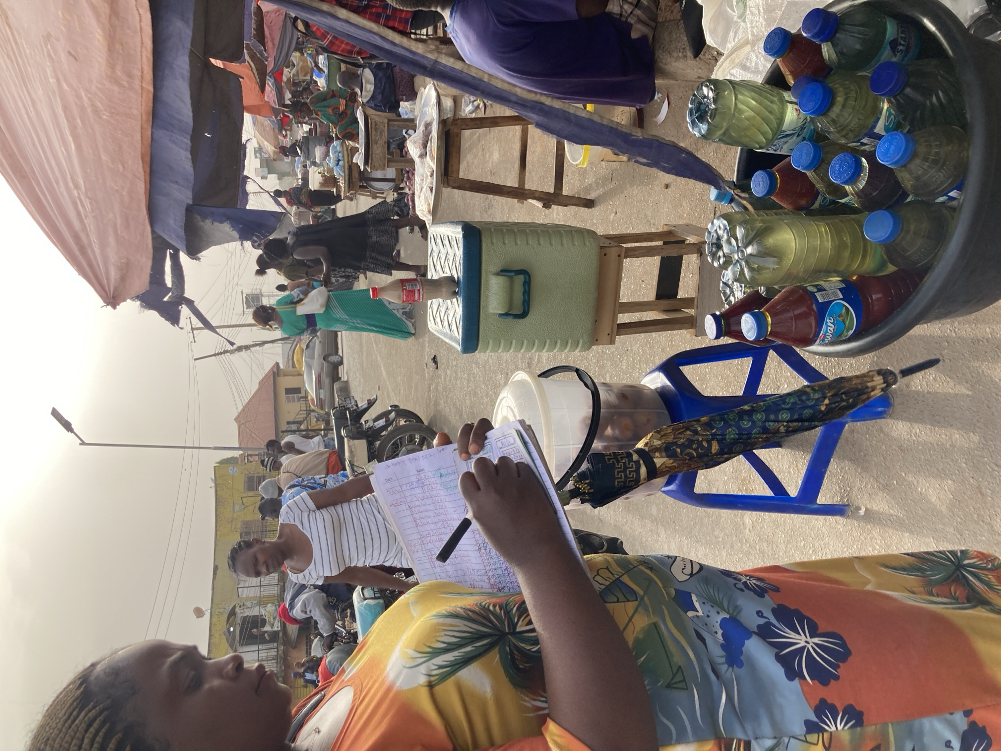 This media file captures a vibrant outdoor market scene in Gwong, Nigeria, during bright daylight hours. The image is taken from a slightly high angle, looking down into the bustling environment.
In the immediate foreground, a person, likely female, with braided hair and wearing a colorful, tropical-patterned top (orange, blue, yellow, green), is seen holding an open notebook and a black pen. They appear to be writing or recording information, possibly sales or attendance, as "DAYS" and "NAME" are visible headings in the notebook, followed by a list of handwritten entries. Another person in a striped shirt stands nearby, looking towards the foreground individual.
To the right, a large blue plastic bucket is filled with numerous plastic bottles containing various liquids, including bottled water (some branded "StarLite") and different colored juices or soft drinks, with "Swan" visible on one red-capped bottle. Next to the bucket is a light green and blue cooler box resting on a wooden stand. A patterned umbrella, featuring blue, gold, and brown designs, leans against a blue plastic chair.
The background reveals a lively street market with various people moving about, some appearing to be vendors behind makeshift stalls with wooden tables and chairs. Tarpaulins and fabric awnings (pink, blue, black) provide shade over market areas. Buildings with yellow and light brown facades are visible further back, along with parked motorcycles and a car. Utility poles and overhead lines crisscross the sky, which is bright and clear, indicating a sunny day. The overall atmosphere suggests a busy, commercial setting.