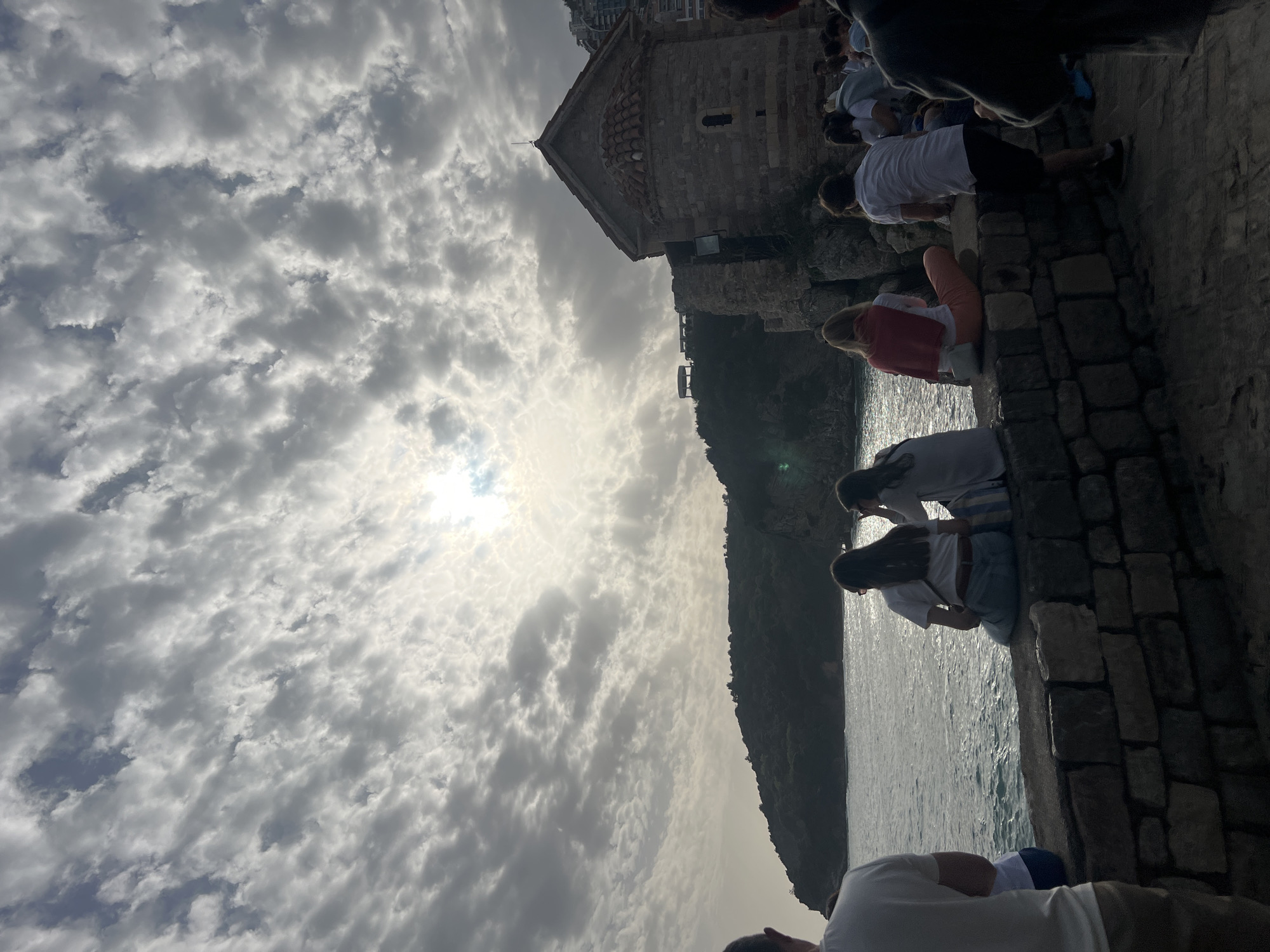 The image captures a view of Sveti Stefan, a small islet and 5-star hotel resort in Montenegro, near Budva. A stone building with a cross on top is visible on the right side of the image. People are sitting and standing on a stone wall. The sea is on the right, and a cloudy sky fills the left side of the frame. The sun is shining through the clouds.