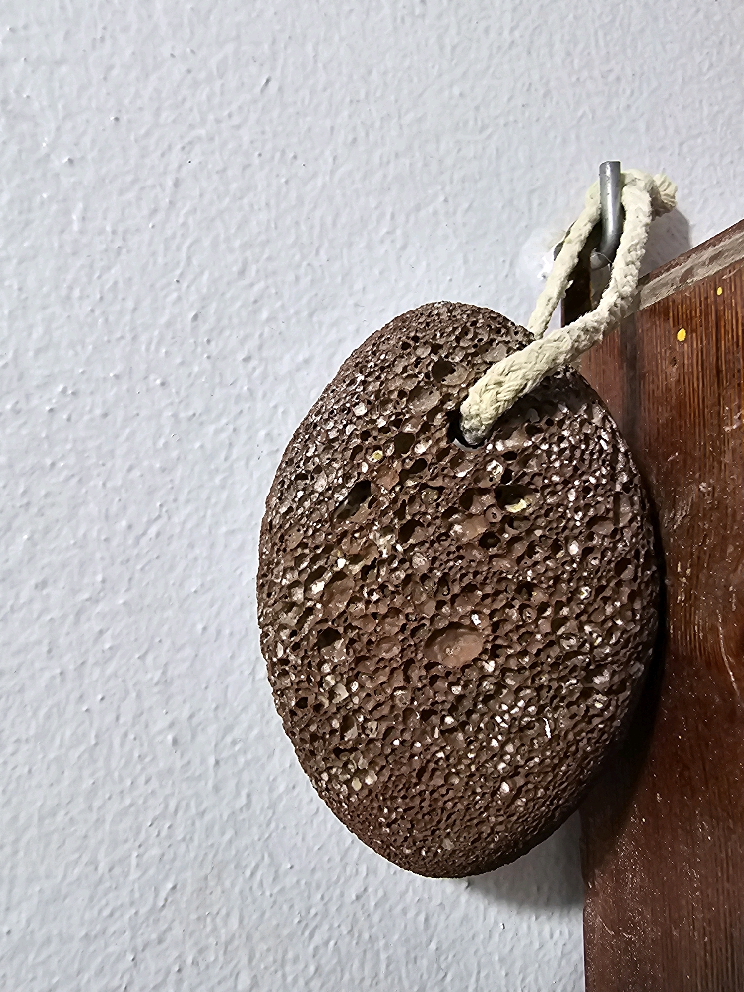 A pumice stone hangs by a rope from a hook attached to a white, textured wall. The pumice stone is oval-shaped and dark brown with numerous pores and a hole through which the rope is threaded. To the right of the pumice stone is a wooden surface, possibly part of a door or cabinet, with a dark brown finish and a few small yellow markings. The image is a close-up view of these objects, with the textured white wall filling the background on the left. The overall scene suggests a utilitarian storage or display in a bathroom or utility area. There is no indication of Batna, Algeria, in the image.