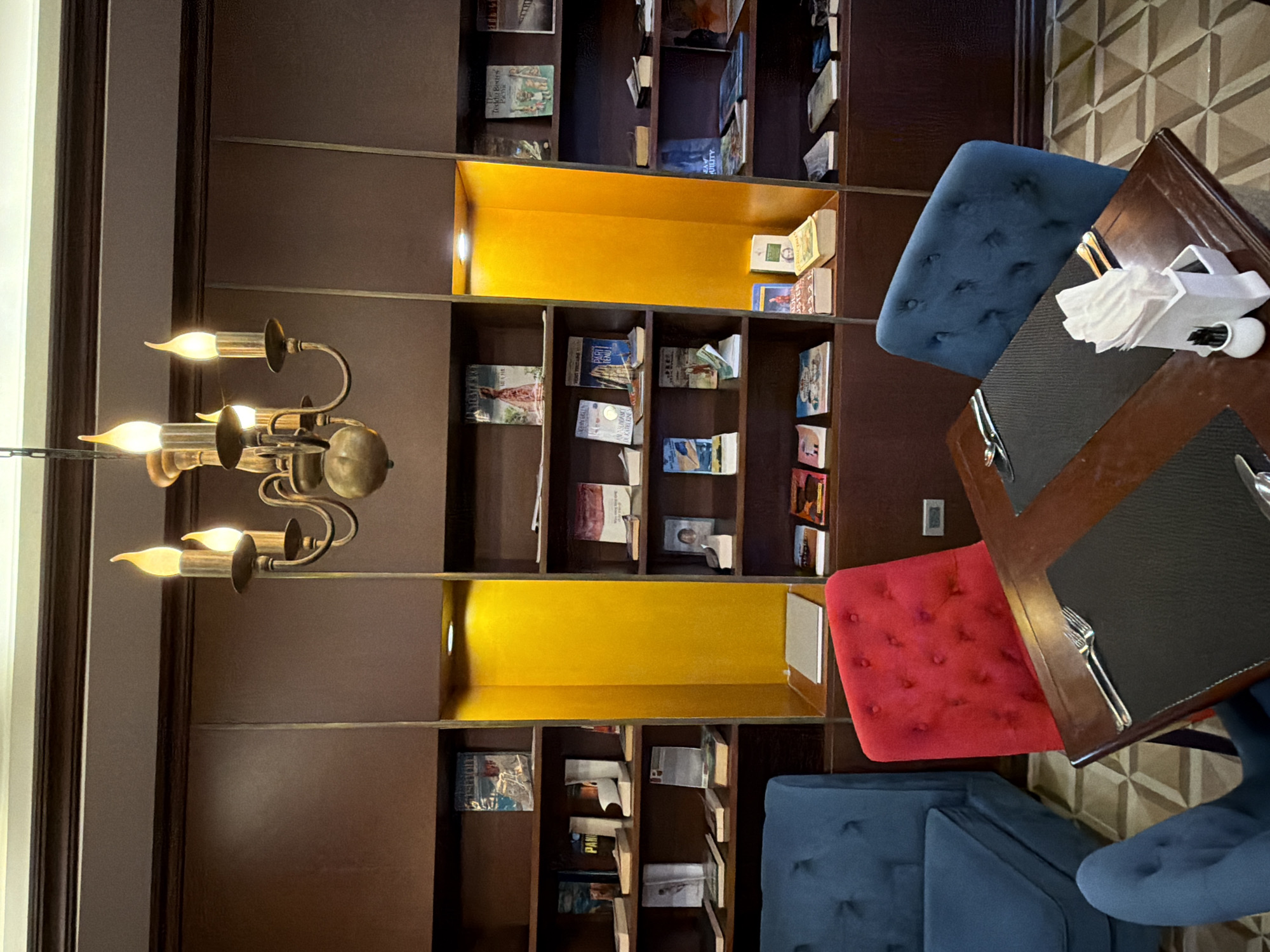 The image depicts the interior of a cafe or restaurant with bookshelves lining a dark wood wall. Several books are displayed on the shelves. In the foreground, a dark wooden table is set with placemats and cutlery. Two chairs, one red and one blue, are visible. In the background, a blue sofa is partially visible. A chandelier hangs from the ceiling, emitting warm light. The floor has a geometric pattern. The cafe appears to be located in Thuận Hóa Ward, Vietnam, though no specific landmarks or individuals are identifiable from the image.