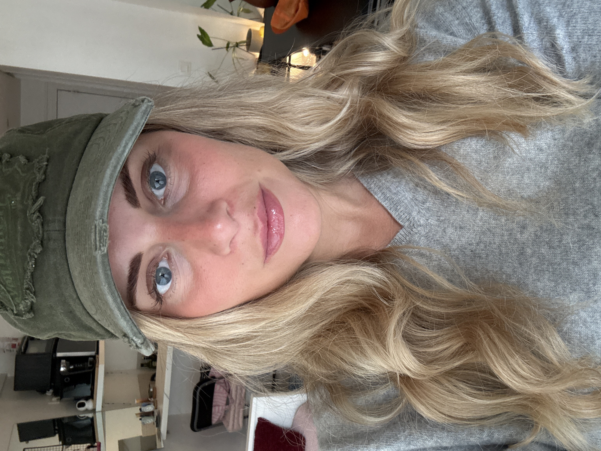 A close-up, slightly rotated, indoor selfie shows a blonde woman with blue eyes and fair skin wearing a green cap and a grey sweater. The background reveals a glimpse of a room with shelves and various objects, suggesting a domestic interior. The specific location in Paris, France, is not identifiable from the image.