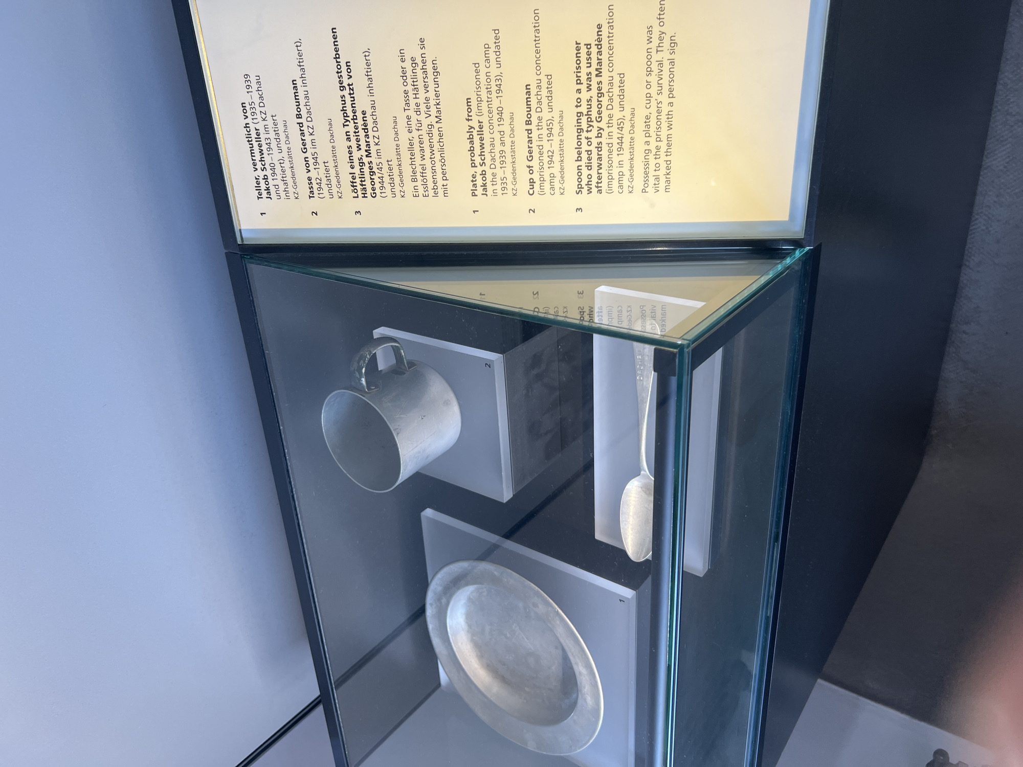 The image shows a display case in the Dachau Concentration Camp Memorial Site in Dachau, Germany. Inside the case are three items: a plate, a cup, and a spoon. A descriptive plaque next to the display case provides information about each item.
The plaque is divided into two columns. The left column is in German and the right column is in English. The German text is as follows:
1. Teller, vermutlich von Jakob Schweller (1935-1939 und 1940-1943 im KZ Dachau inhaftiert), undatiert. KZ-Gedenkstätte Dachau.
2. Tasse von Gerard Bouman (1942-1945 im KZ Dachau inhaftiert), undatiert. KZ-Gedenkstätte Dachau.
3. Löffel eines an Tyhus gestorbenen Häftlings, weiterbenutzt von Georges Maradène (1944/45 im KZ Dachau inhaftiert), undatiert. KZ-Gedenkstätte Dachau.
Ein Blechteller, eine Tasse oder ein Esslöffel waren für die Häftlinge lebensnotwendig. Viele versahen sie mit persönlichen Markierungen.
The English text is as follows:
1. Plate, probably from Jakob Schweller (imprisoned in the Dachau concentration camp 1935-1939 and 1940-1943), undated. KZ-Gedenkstätte Dachau.
2. Cup of Gerard Bouman (imprisoned in the Dachau concentration camp 1942-1945), undated. KZ-Gedenkstätte Dachau.
3. Spoon belonging to a prisoner who died of typhus, was used afterwards by Georges Maradène (imprisoned in the Dachau concentration camp in 1944/45), undated. KZ-Gedenkstätte Dachau.
Possessing a plate, cup or spoon was vital to the prisoners' survival. They often marked them with a personal sign.
The plate is a simple, shallow metal dish. The cup is a cylindrical metal mug with a handle. The spoon is a standard metal spoon. All three items appear to be made of aluminum or a similar lightweight metal. They are displayed on white risers inside a glass-fronted display case. The lighting in the display case is consistent, illuminating the artifacts without harsh shadows. The background of the display case is dark, helping to highlight the items. The plaque is positioned to the right of the display case and is illuminated by ambient light. The overall environment suggests a museum or memorial exhibition.