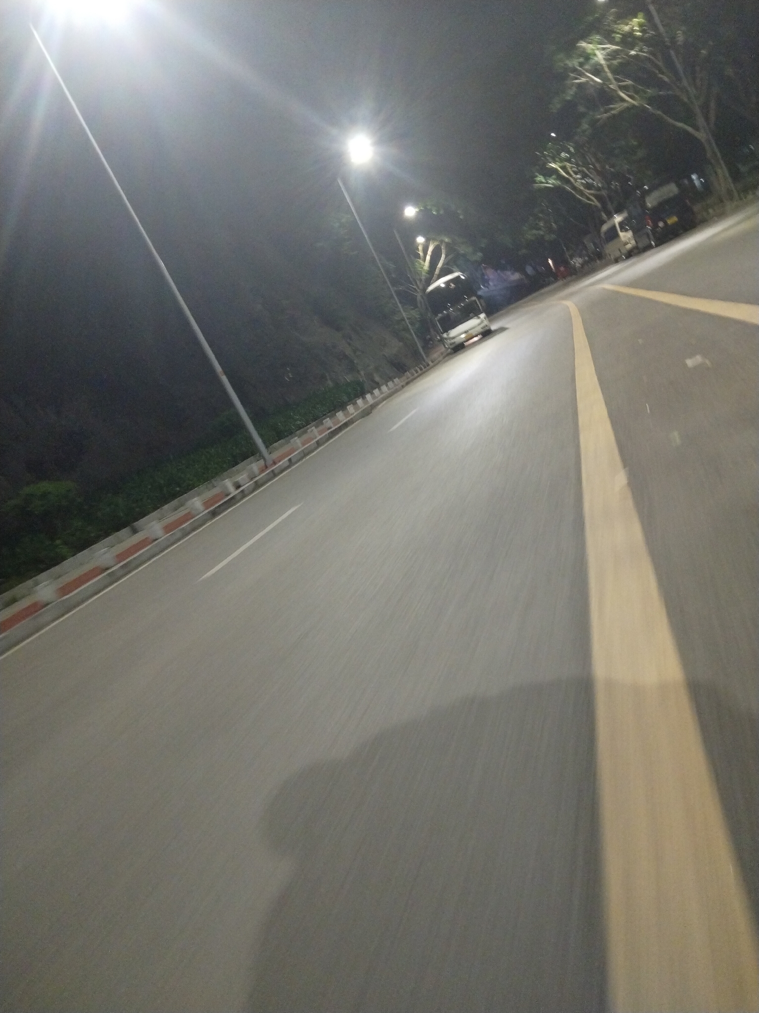 The image depicts a night scene on a road in Jiulongpo District, China. The road is well-lit by streetlights, and a white vehicle is visible in the distance. There is a yellow line indicating the lane separation on the right side of the road. The road surface appears smooth and grey. On the left side of the road, there is a curb with a red and white pattern, followed by vegetation. The overall environment suggests an urban or suburban setting at night.