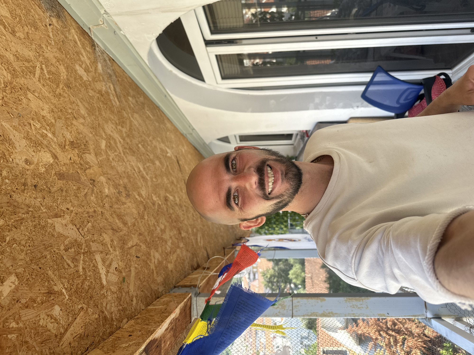 A smiling, bald man with a beard stands on a balcony, likely in Budva, Montenegro. He's wearing a light-colored shirt and a gold necklace. Behind him, a string of colorful prayer flags hangs along the balcony railing. The balcony overlooks a residential area with rooftops and trees. To the left, there's a wall constructed of OSB. Above, a window with an arched top is visible. A blue seat cushion and a red and black object sit on the windowsill.