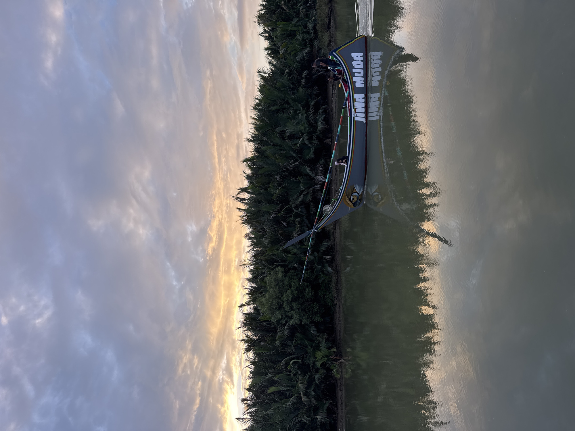 At sunset in Rhieng Mancang, Indonesia, a gray boat with the words "JIWA MUDA" painted in white lettering travels down a calm river. The boat has a stylized eye painted on its bow and a multi-colored stripe along its side. Several individuals are on the boat, and one appears to be steering. Dense foliage, likely palm trees, lines the riverbank, reflecting on the water's surface along with the boat and the colorful sky. The sky displays hues of orange, yellow, and purple from the setting sun, with scattered clouds. The water is a muted green color.