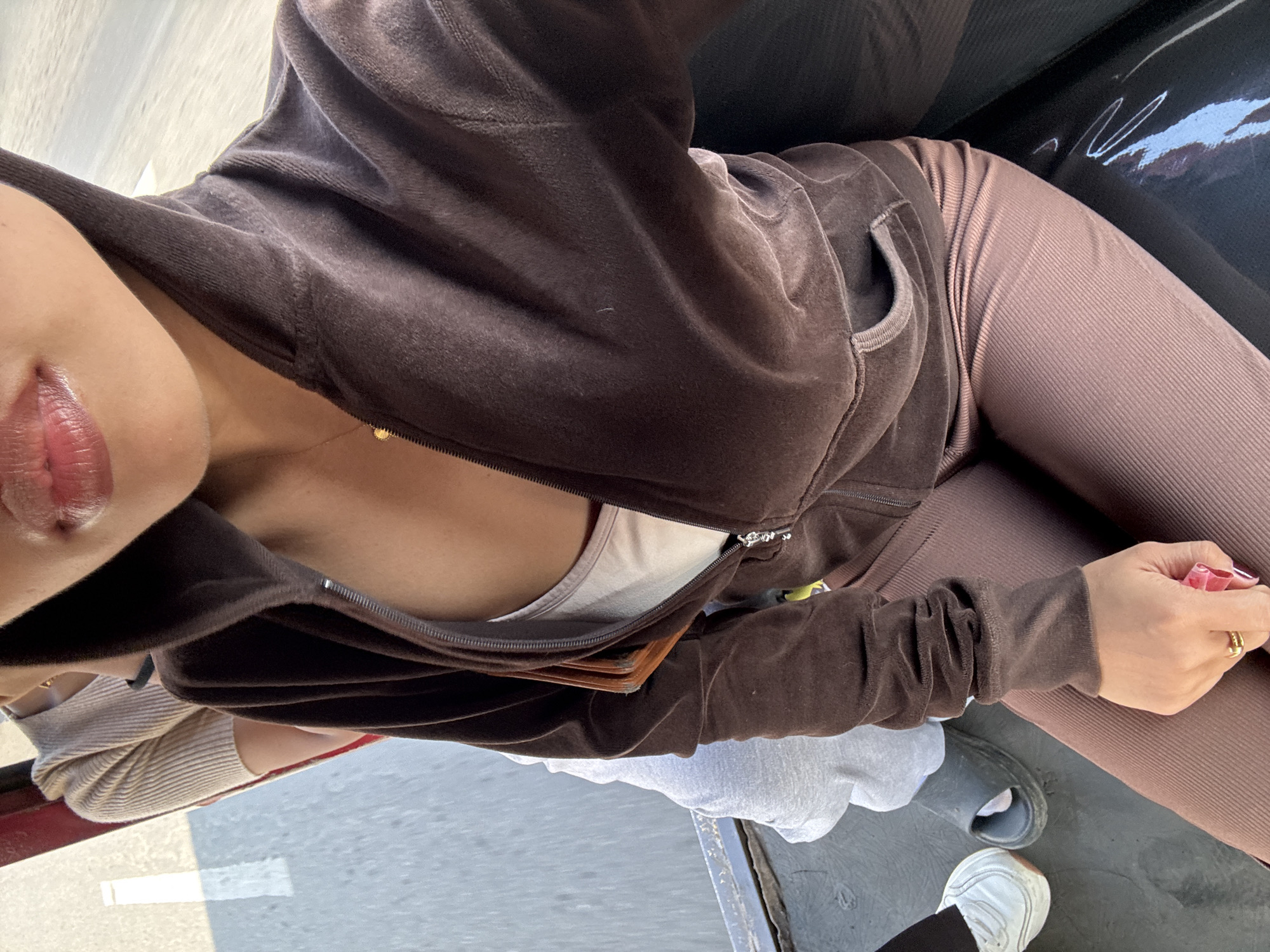 The image shows a close-up of a person from the chest up, sitting in what appears to be a trishaw, a common mode of transportation in the Philippines, including Davao City. The person is wearing a brown velour zip-up hoodie, partially unzipped to reveal a light-colored tank top. They are also wearing tight, ribbed, mauve-colored pants. The person's hands are visible, with one resting on their lap, adorned with a gold ring. They are holding a small pink object in their hand. In the background, another person is partially visible, wearing a beige ribbed top and grey pants. The ground outside the vehicle appears to be a paved road, suggesting an urban environment, consistent with Davao City. The lighting indicates it is daytime, with sunlight casting shadows.