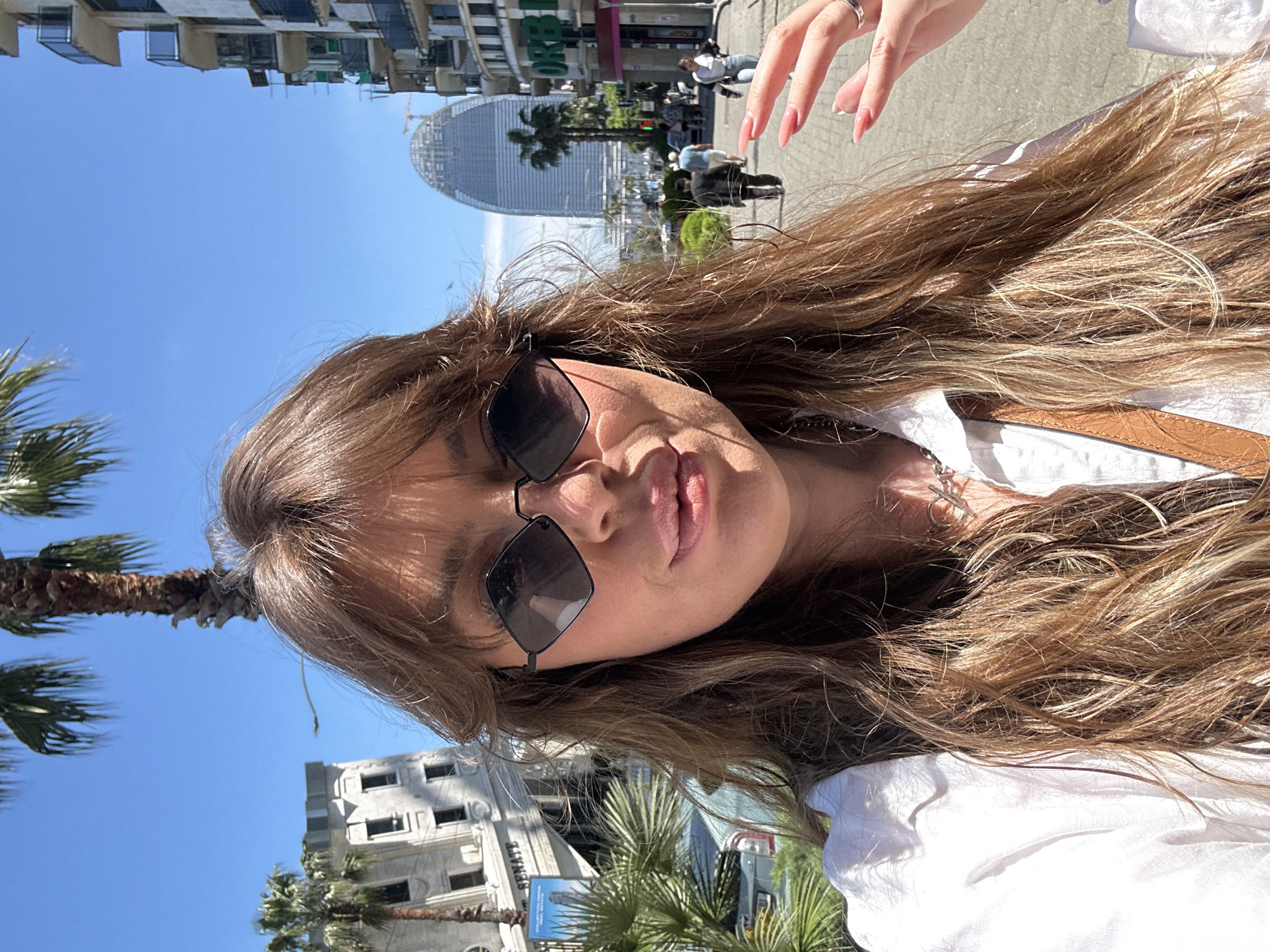 A selfie-style photograph shows a woman with long brown hair, wearing sunglasses and a white shirt. She is outdoors in Batumi, Georgia, with a building that appears to be the Radisson Blu Hotel Batumi visible in the background. The woman is facing the camera, with a slight upward angle. The background shows a blue sky and palm trees. Other buildings and pedestrian traffic are visible further in the background.
