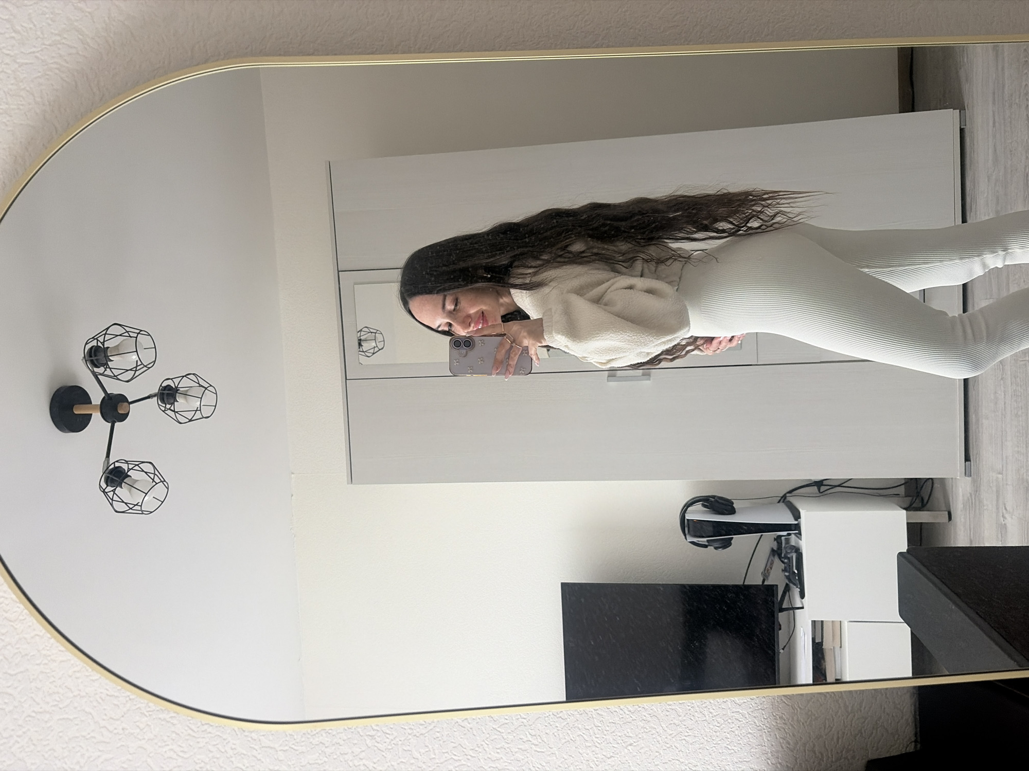 A medium shot shows a woman reflected in a gold-framed mirror. The woman is holding a mobile phone, presumably taking the picture. She is wearing a white, ribbed, fitted two-piece outfit and a cream-colored sweater. Her dark hair is long and flowing. Behind her in the reflection are a white wardrobe, a black television, a white shelf with a gaming console, and part of a black speaker. On the wall to the left, also in the reflection, is a black modern light fixture. The wallpaper is textured and appears to be cream or off-white. The location is likely an indoor setting, and there is no direct evidence to identify it as being in Saint Petersburg, Russia.