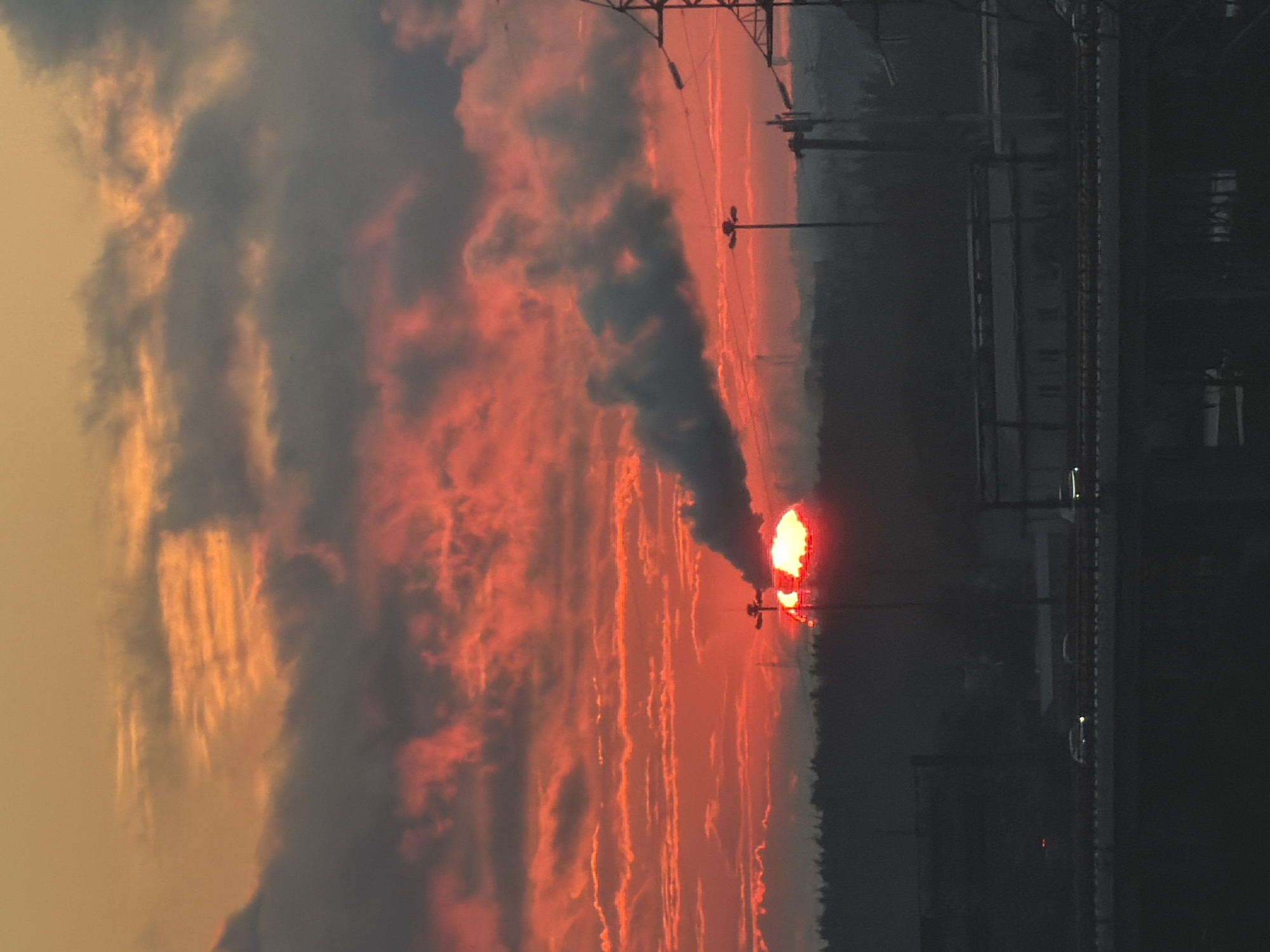 A dramatic sunset paints the sky with vibrant oranges and reds, set against a backdrop of dark, brooding clouds. The sun, a fiery orb, is partially obscured by a plume of grey smoke emanating from what appears to be an industrial facility. In the foreground, silhouetted against the dramatic sky, is a dense line of trees, suggesting a natural border to an urban landscape. A highway stretches across the midground, with a few vehicles visible on its surface, their headlights casting faint glows. In the distance, structures and power lines hint at the presence of a city, their details softened by the atmospheric haze of dusk. The overall impression is one of industrial power meeting the raw beauty of nature.