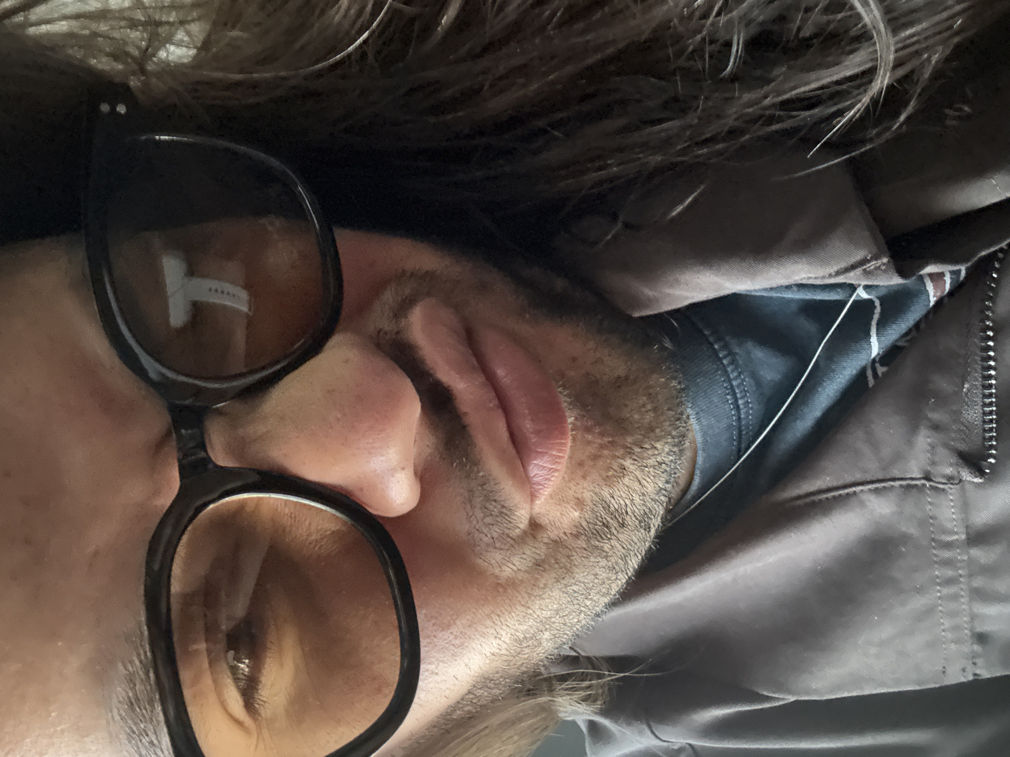 A close-up, horizontally oriented shot captures the left side of a person's face, head tilted slightly to the right. The individual, appearing to be an adult male, wears dark-framed sunglasses with amber-tinted lenses. Through the left lens, a partially visible eye can be seen. A prominent reflection in the left sunglass lens reveals a white T-shaped object, possibly a stylized logo or part of a digital display, with small circular dots aligned vertically next to its right side.
The person has medium brown hair, which appears somewhat long and is visible cascading around the head and ear. Short, dark stubble covers the cheek and jawline. The ear, lips, and part of the nose are in view.
The individual is wearing dark clothing: a dark blue or gray shirt is visible beneath a dark jacket, possibly gray or dark brown, with a silver zipper on the right side. A thin white string or thread is noticeable against the dark fabric of the jacket and shirt. The overall impression is neutral and relaxed. The lighting suggests natural illumination. The image context is Valle de Chalco Solidaridad, Mexico.