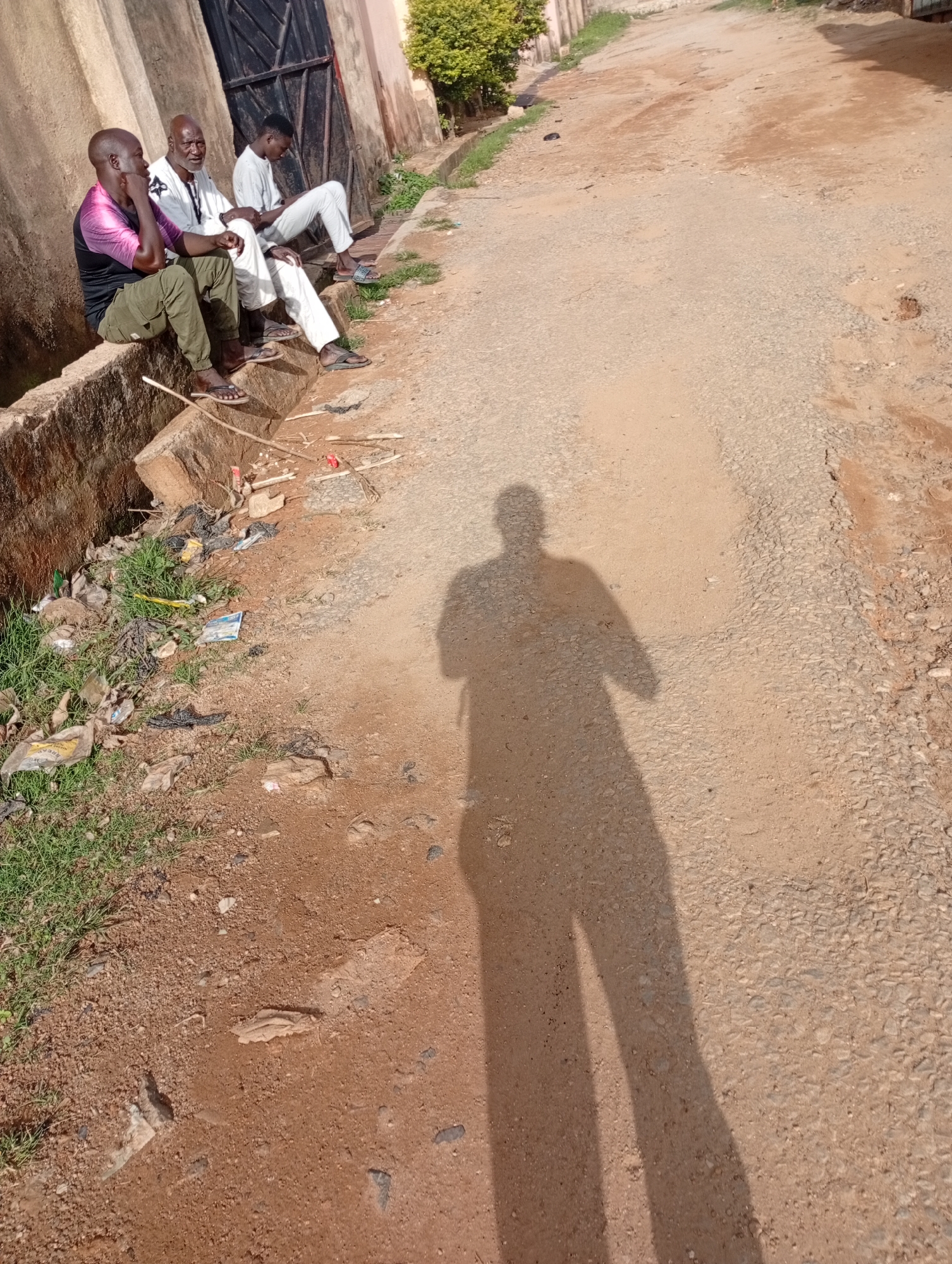In Rafin Par, Nigeria, three men are seated on a concrete ledge next to a dirt road. The man on the left wears a pink and black patterned shirt and green trousers. The man in the middle is older, with a grey beard, and wears a white traditional outfit. The man on the right is younger and wears a white outfit. They appear to be engaged in conversation or simply resting. The surrounding environment is dusty with some overgrown grass and scattered debris along the edge of the road. The sun casts a long shadow of the photographer onto the road, indicating it is likely daytime.