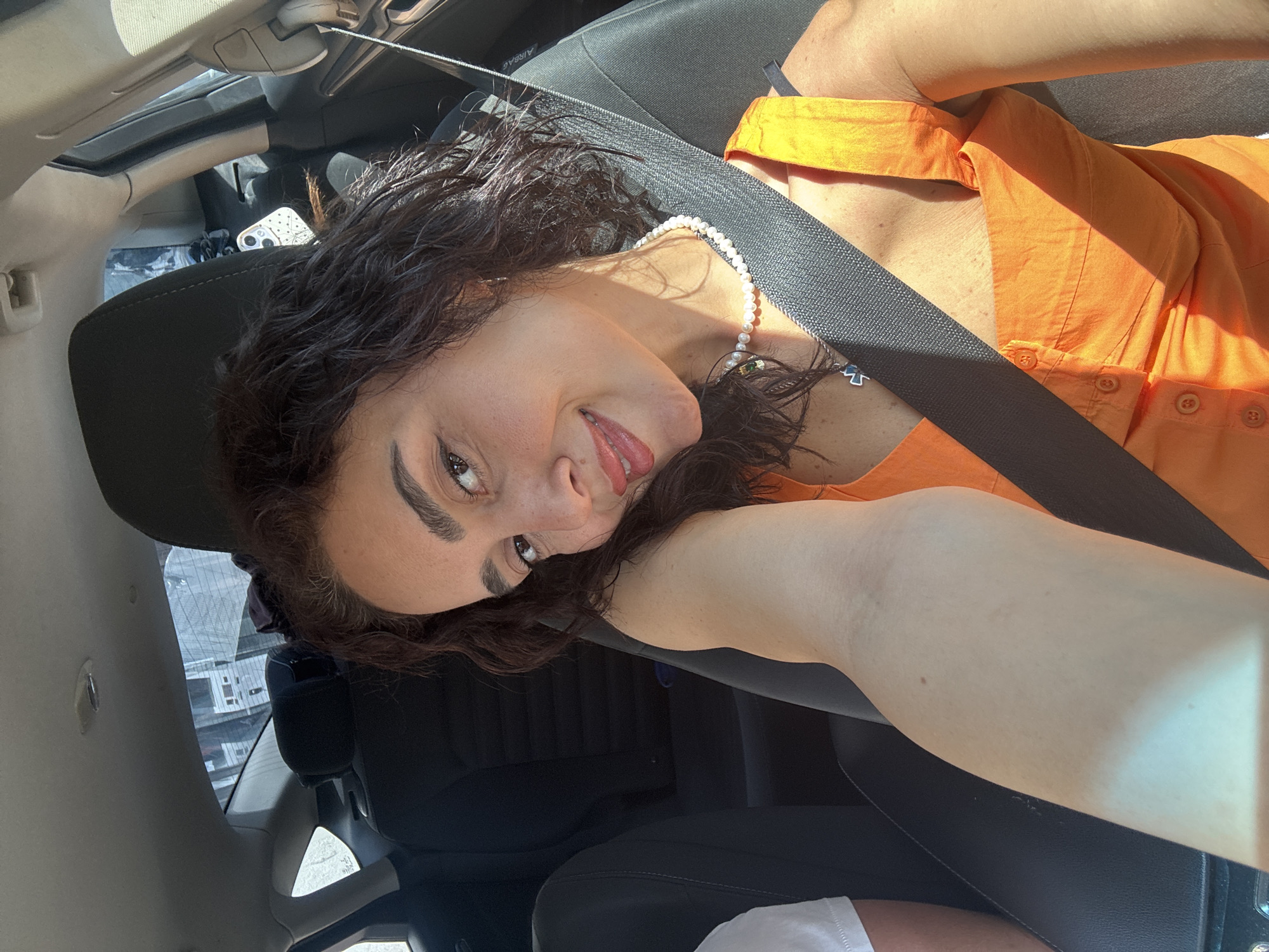 A woman with curly dark brown hair and light brown skin is taking a selfie in the passenger seat of a car. She is wearing an orange sleeveless top and a pearl necklace. Her right arm is bent, with her hand resting on her head. She is smiling and looking at the camera. The background shows the interior of the car, including black seats and a seatbelt. Sunlight streams through the windows, casting shadows on the woman and the car's interior. The location is not identifiable as Tbilisi, Georgia.
