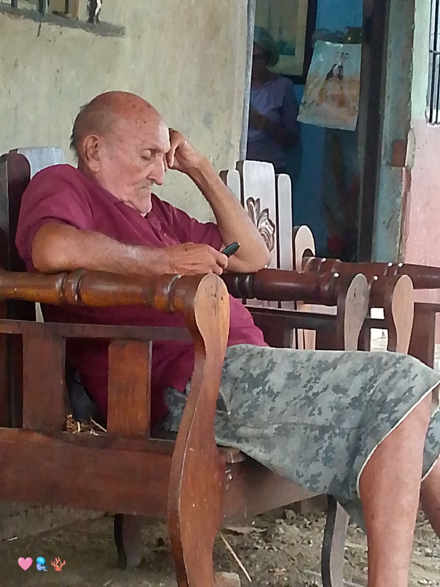 An elderly bald man with olive skin and wrinkles is seated in a wooden rocking chair, looking down and resting his head on his hand. He is wearing a maroon t-shirt and camouflage shorts. The man is holding a black mobile phone in his right hand. The rocking chair is made of dark wood with carved details. In the background, a woman in a purple shirt is visible through an open doorway, with a framed picture hanging on the wall. The scene appears to be an outdoor setting, possibly a patio or veranda, with dirt and debris on the ground. The overall impression is one of quiet repose. The scene is located in San Sebastián, Venezuela.
