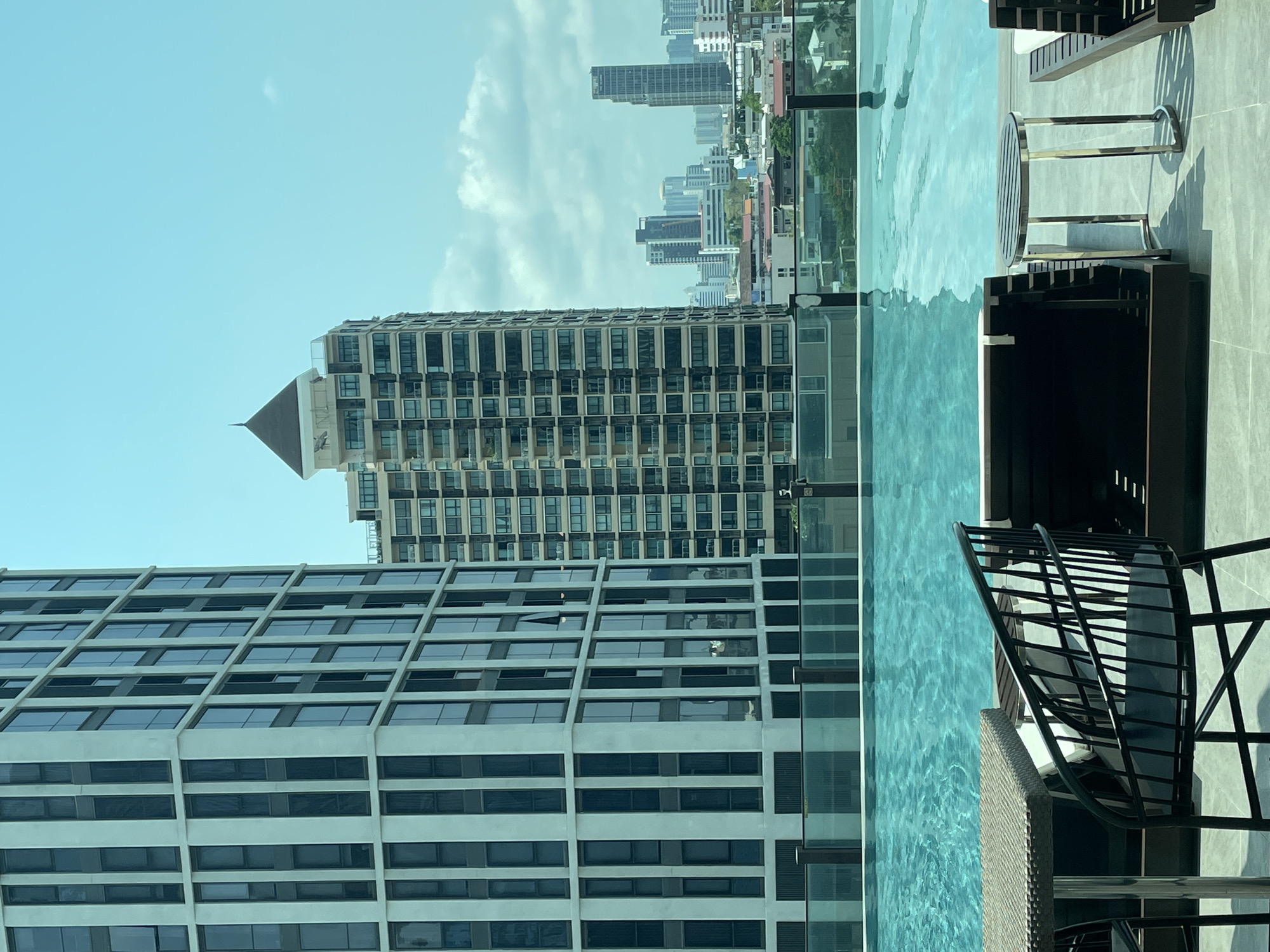 The image displays a serene rooftop infinity pool setting overlooking a dense urban landscape, identified as Bangkok, Thailand. In the foreground, the inviting, clear turquoise water of the infinity pool occupies the lower portion of the frame, reflecting the bright, mostly clear blue sky with scattered white clouds. To the right of the pool, a paved patio area is furnished with dark, modern outdoor chairs or loungers and a small, round table with a metallic base, casting crisp shadows that indicate it is daytime, likely late morning or early afternoon.
Beyond the pool, a panorama of tall, modern buildings defines the city skyline. A prominent light-colored building with a distinctive grid pattern of windows takes up a large portion of the mid-ground on the left. Further back and slightly to its right, another multi-story building features a unique dark, pyramid-shaped roof structure. The distant background reveals a sprawling cityscape filled with numerous high-rise buildings stretching towards the horizon. Faint text, possibly "ELITE," is barely discernible on one of the distant buildings. No people are visible, contributing to the tranquil and expansive atmosphere of this luxurious urban retreat.