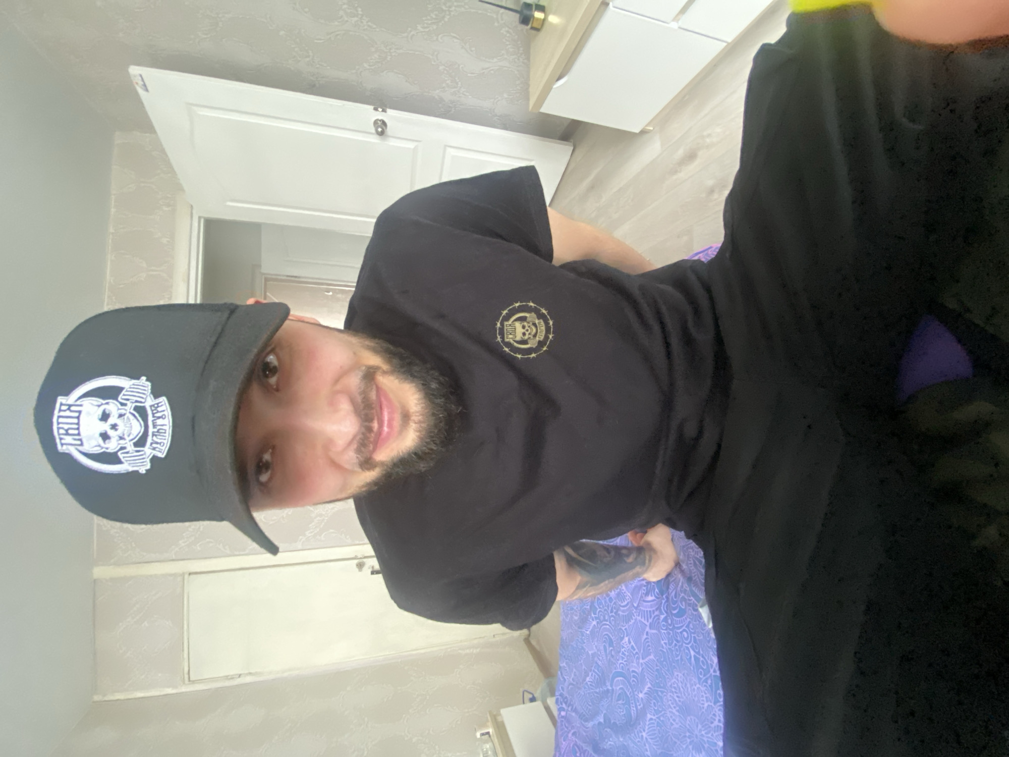 A bearded man wearing a black t-shirt and cap is indoors, likely in an apartment in Chaikovsky, Russia. The man is partially visible from the waist up. He is sitting or lying on a patterned blue and purple surface. The background includes a white door and a dresser. The man has a tattoo on his left arm.