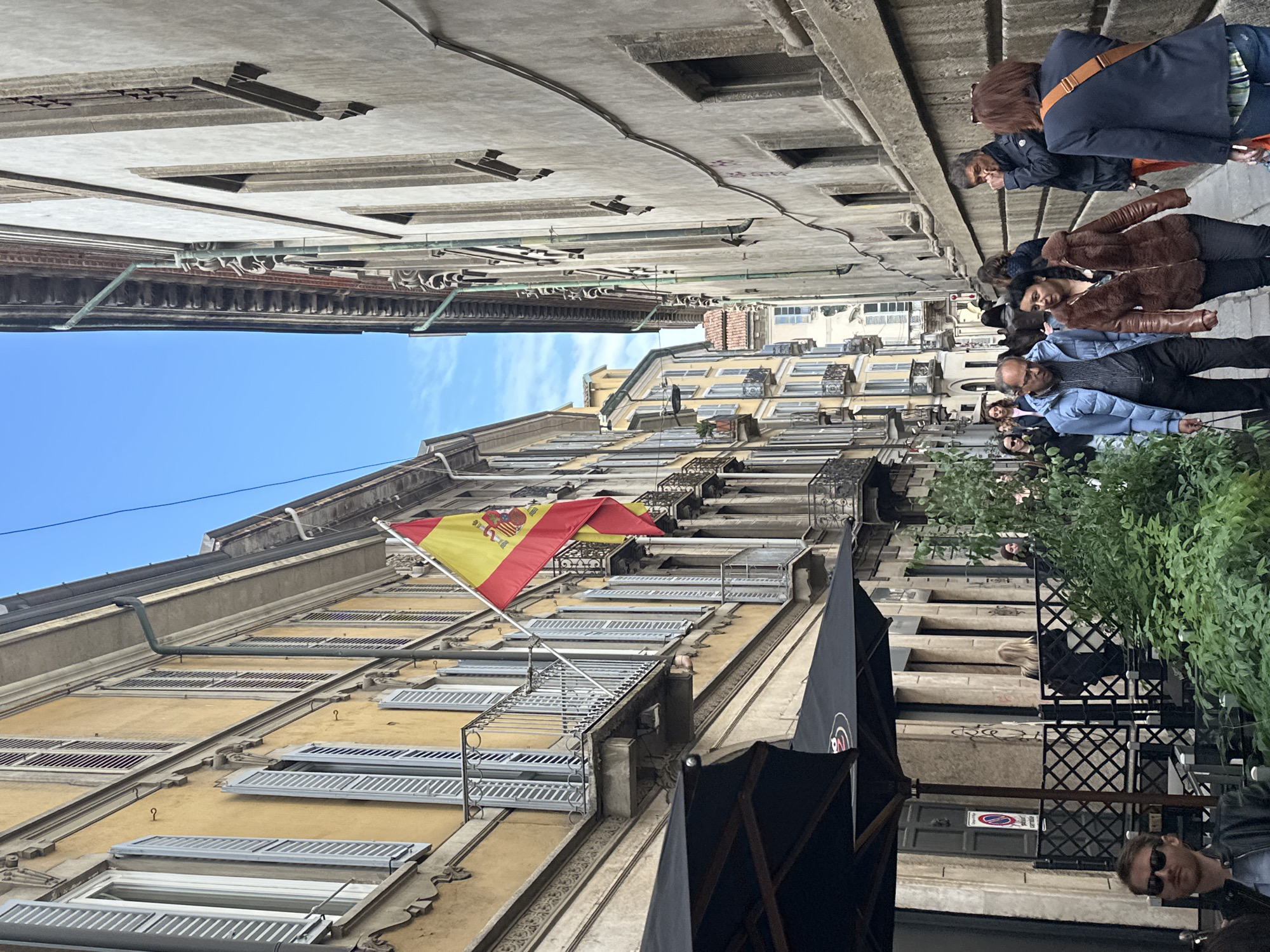 The image captures a lively street scene in Milan, Italy, featuring a narrow thoroughfare flanked by tall, multi-story buildings under a clear blue sky. The perspective is looking up a street, with people populating the foreground and middle ground.
On the left side, a prominent building with a light-colored facade displays numerous windows with louvered shutters and ornate balconies. A Spanish flag, featuring the red and yellow stripes and the Spanish coat of arms, is proudly flown from one of its balconies. Exposed pipes and wiring run along the building's exterior.
The street level on the right appears to host an outdoor cafe or restaurant, indicated by black canopies, tables, and chairs. Several individuals are seated or standing in this area, some casually walking, others perhaps engaging in conversation. People of various ages are present, dressed in casual attire. Notable individuals include a man in sunglasses partially visible in the bottom right, and a woman in a brown fur-collared jacket among a group standing.
The architecture of the buildings is characteristic of European cities, with detailed cornices, balconies, and traditional window treatments. Some building facades show signs of aging, with weathered surfaces and visible utility installations. In the distance, more buildings line the street.
The time of day is daytime, evidenced by the bright, natural light and the clear blue sky. The weather appears pleasant. A red and white circular street sign, possibly indicating a restriction, is partially visible on a building across the street from the cafe area.
