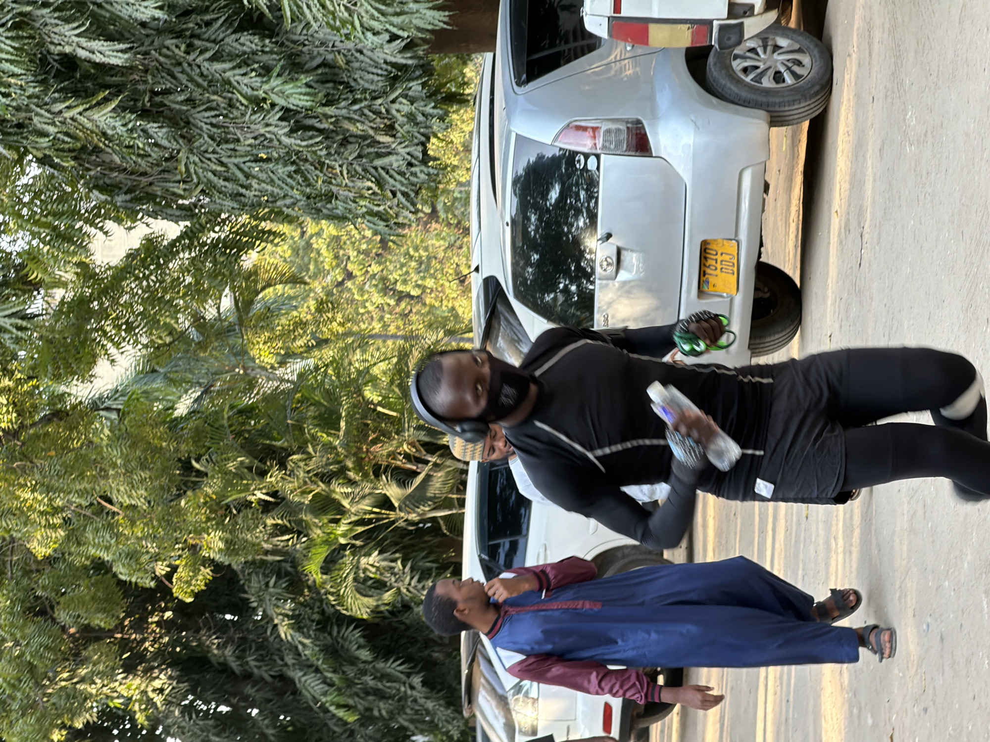 In Dar es Salaam, Tanzania, a man wearing a black long-sleeved shirt, black shorts, black leggings, headphones, and a black face mask runs forward, holding a water bottle and a green item. He is in the foreground, slightly to the right of the frame. Behind him, a man in a blue and maroon thobe with sandals walks to the left, looking to his right. A third person, wearing a white shirt and a patterned cap, stands between them, partially obscured. Several parked cars are visible, including a silver hatchback with a yellow license plate reading "T610 DDJ". The environment appears to be an outdoor, urban setting with trees and sunlight casting shadows.