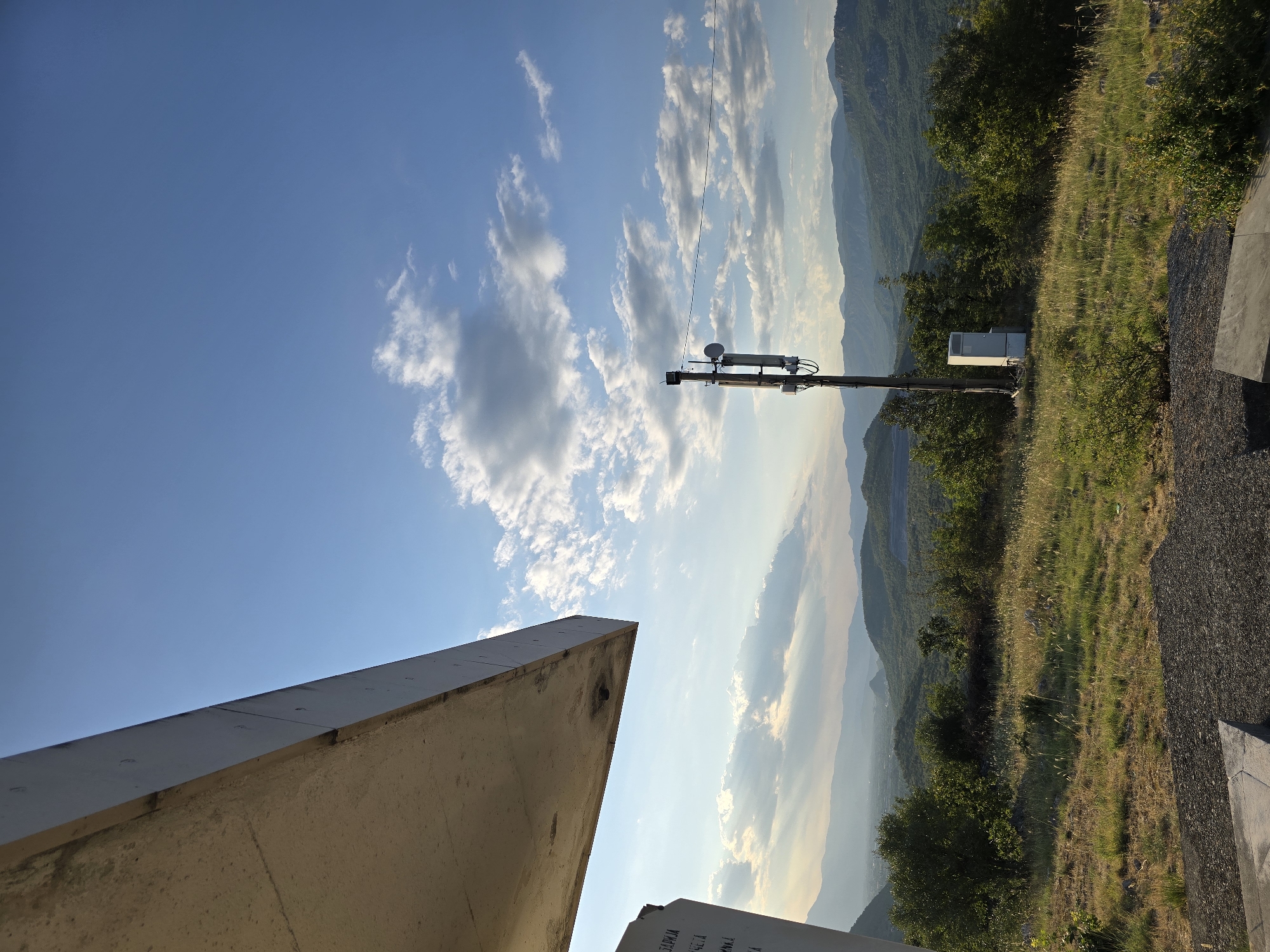 The image depicts an outdoor scene with a vast landscape under a clear blue sky with scattered clouds. In the foreground, a concrete structure, possibly part of a building or monument, extends from the left side of the frame, casting a shadow. To the right of the concrete structure, a grassy hill is visible with dry grass and some green bushes. A utility pole with antennas and a small white cabinet at its base stands prominently on the hill. Beyond the immediate foreground, the landscape unfolds into rolling hills covered with lush green vegetation and trees. In the distance, a range of mountains stretches across the horizon, bathed in the soft light of what appears to be either sunrise or sunset. The overall atmosphere is serene and natural, with a touch of man-made infrastructure. There are no people or animals visible in the image.