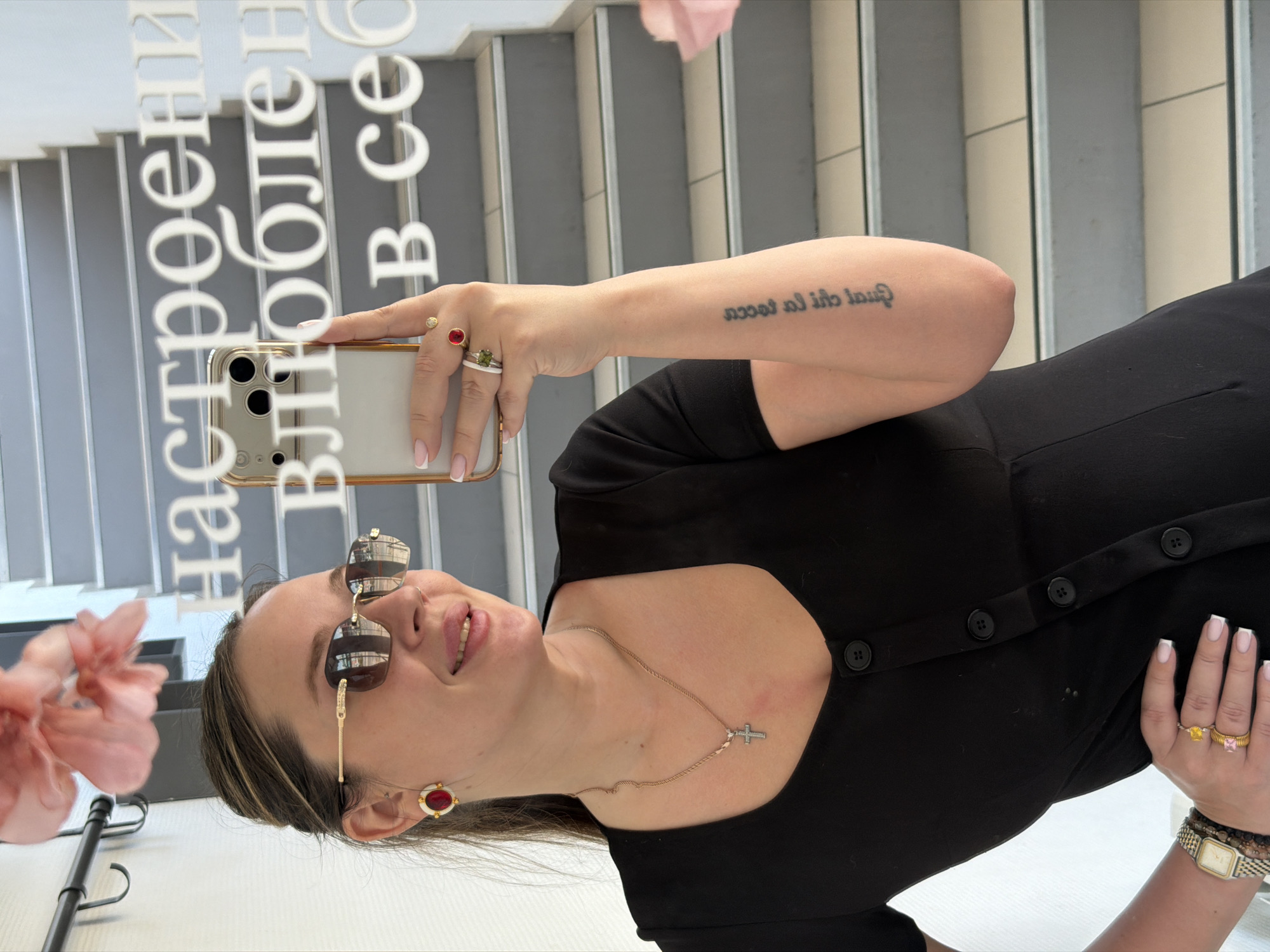 A woman is taking a selfie indoors. She wears a black dress, sunglasses, and jewelry, including a necklace with a cross pendant, rings, a watch, and earrings. She is holding a gold-colored smartphone with three rear-facing cameras. The background shows a staircase and a wall with Russian text that reads "настроение в сезоне" (mood in season). The image appears to be taken in the city of Chaikovsky, Russia. A pink flower is visible to the left.