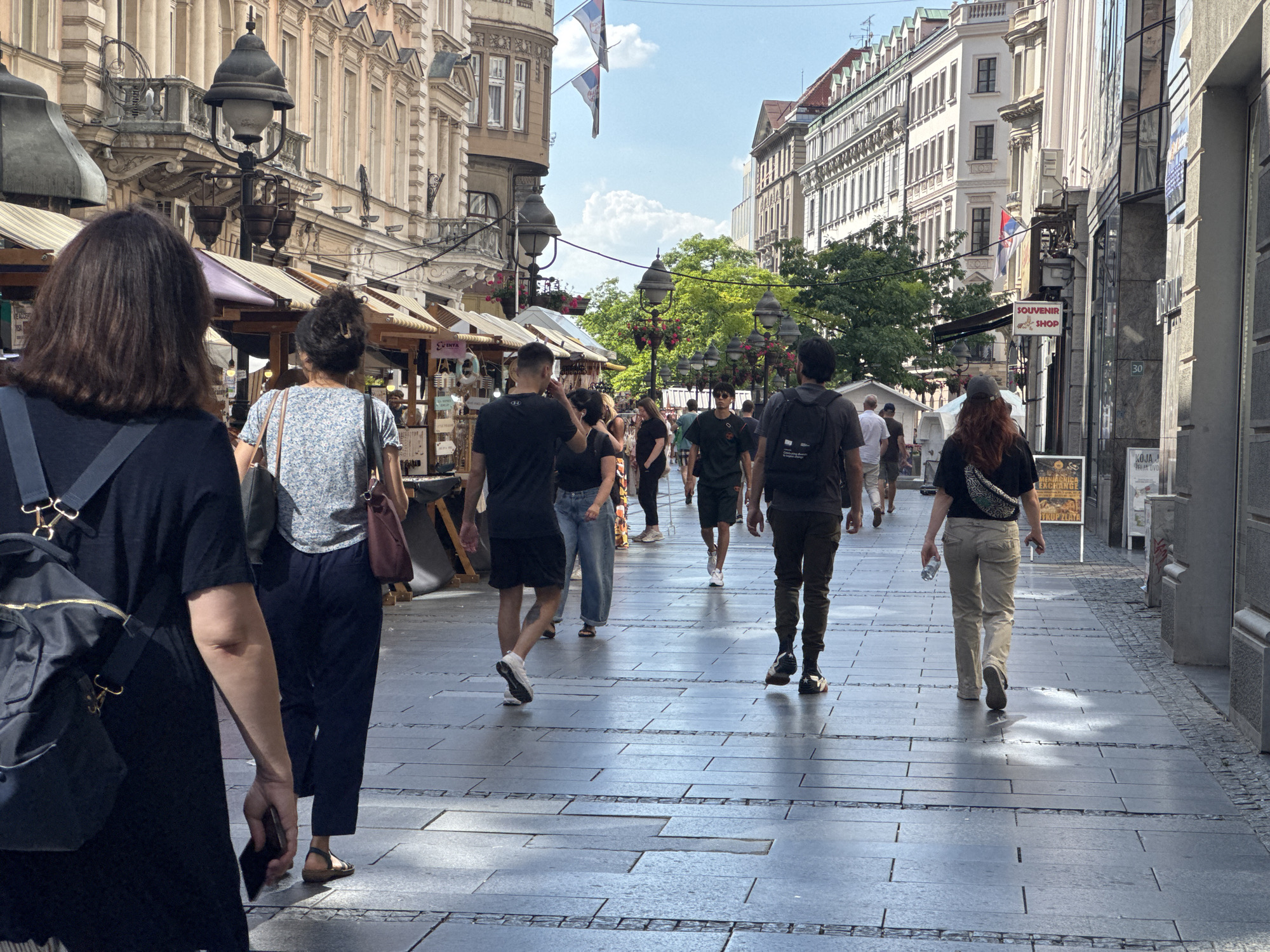 The image shows a street scene in Belgrade, Serbia, with several people walking along a paved sidewalk lined with buildings and market stalls. The day appears to be sunny and warm, casting shadows on the ground.
In the foreground, a woman with dark hair seen from behind is walking away from the camera, carrying a black backpack. To her right, another woman in a floral-patterned shirt and dark pants is also walking forward. Several other individuals, including men and women, are visible walking in the same direction, some carrying bags and wearing casual attire. The crowd suggests a bustling public space.
The buildings on either side of the street are multi-story and feature architectural details typical of European city centers, with ornate facades and balconies. Some buildings have awnings or umbrellas indicating shops or market stalls. Flags, possibly of Serbia, are visible on one of the buildings. There are also street lamps lining the walkway, adding to the urban ambiance.
A sign for a "SOUVENIR SHOP" is clearly visible on the right side of the street. Other text is present on signs and posters, but not readily legible. The overall mood is one of a lively, everyday scene in a European city, likely during daytime given the bright sunlight and clear sky with some clouds.