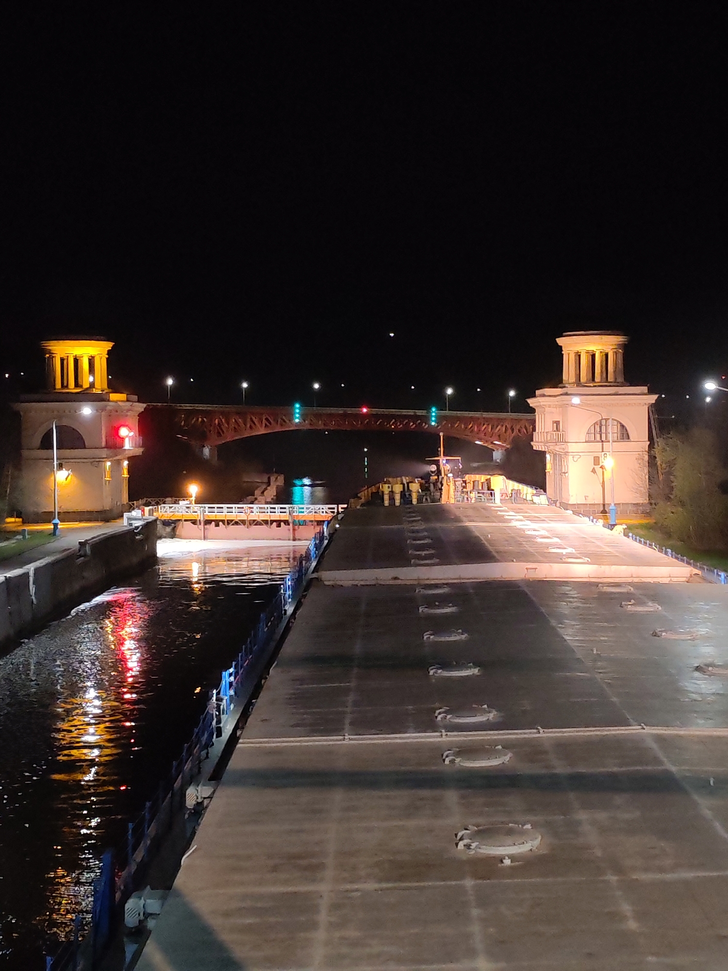 At night in Iksha, Russia, a wide, concrete waterway extends from the foreground, bordered by a blue railing on the left. The water reflects red and yellow light from various sources. On either side of the waterway, structures with illuminated, pillared towers stand. Beyond these structures, a red truss bridge spans the waterway, showing red and green lights. In the center of the waterway, a floating platform is present, occupied by individuals in yellow clothing, likely construction workers. The sky is dark, and there are scattered lights in the distance.