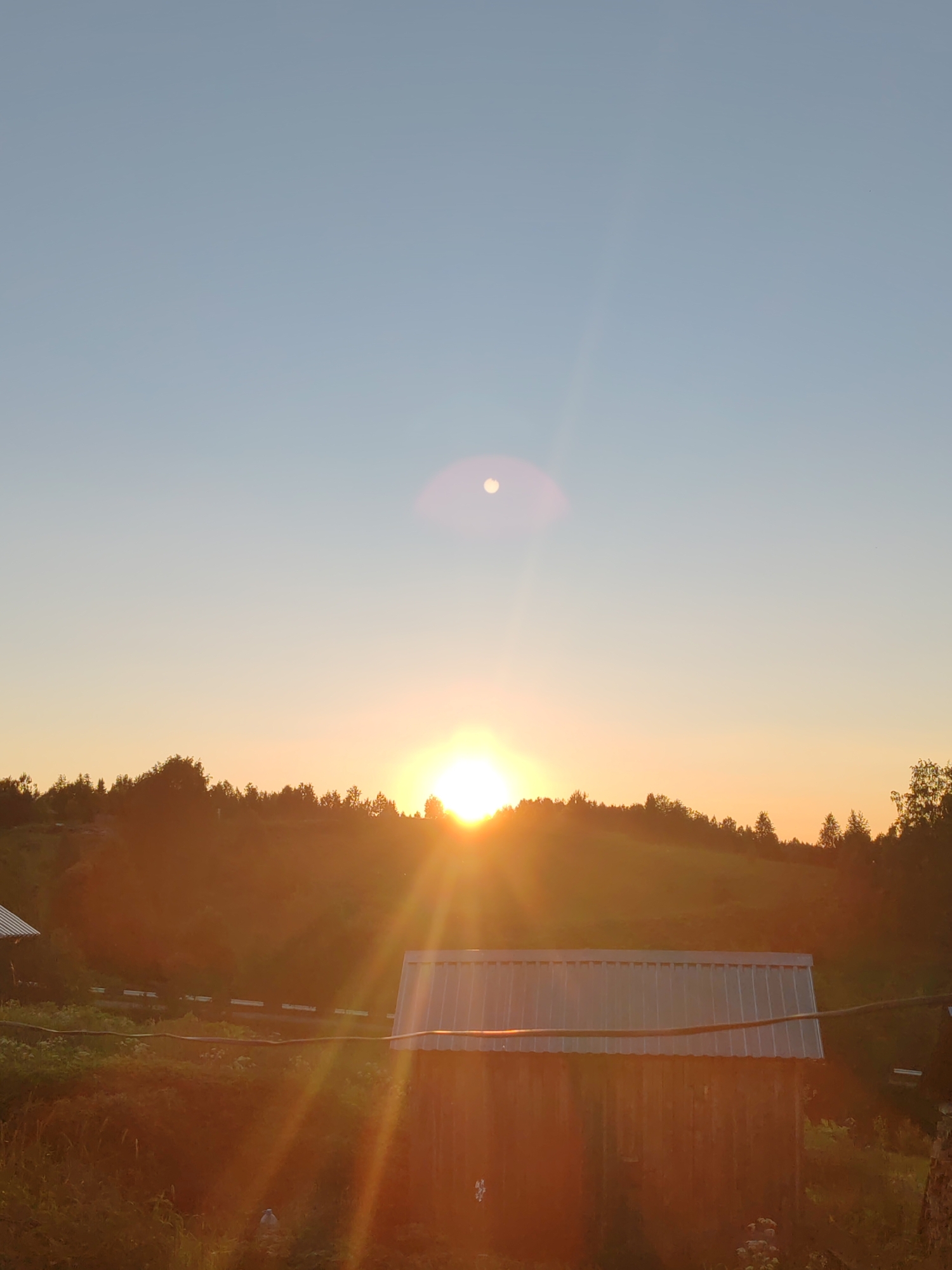 The image depicts a rural landscape at sunset. The sky is a gradient of pale blue at the top transitioning to a warm orange and yellow near the horizon, indicating the setting sun. The sun itself is visible as a bright, glowing orb just above a tree-lined ridge, casting strong rays of light that create lens flare and illuminate the scene. A hazy, circular lens flare is also present higher in the sky.
In the foreground, a rustic wooden building with a corrugated metal roof is situated. The building appears weathered, with visible wood grain and vertical planks. To the left of this building, partially obscured, is another structure with a similar metal roof. The ground around the buildings is covered in vegetation, appearing somewhat overgrown with grasses and other plants. A thin, dark cable runs horizontally across the image in front of the building's roof. In the background, a rolling hill covered with dense trees forms the horizon line, silhouetted against the bright sunset. The overall atmosphere is serene and evokes a sense of twilight in a natural setting. The location is identified as Чухлэм, Russia.