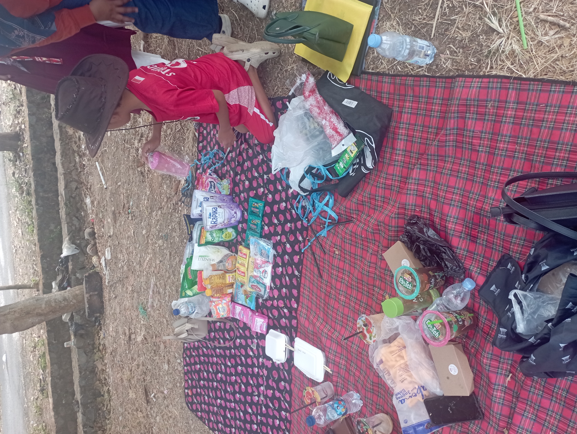 The image depicts a picnic scene with various food and drink items spread across two picnic blankets on the ground, likely in an outdoor park or recreational area in Purwakarta, Indonesia, given the context. A young person wearing a red t-shirt and a brown cowboy hat is kneeling on a blanket, holding a pink cup. Other individuals are partially visible in the background. The picnic spread includes bottled water, juice boxes, snacks like biscuits and packaged treats, and possibly some prepared food in containers. The ground is covered with dry leaves and straw, with a concrete embankment and a road visible in the upper portion of the frame. The overall atmosphere suggests a casual outdoor gathering.