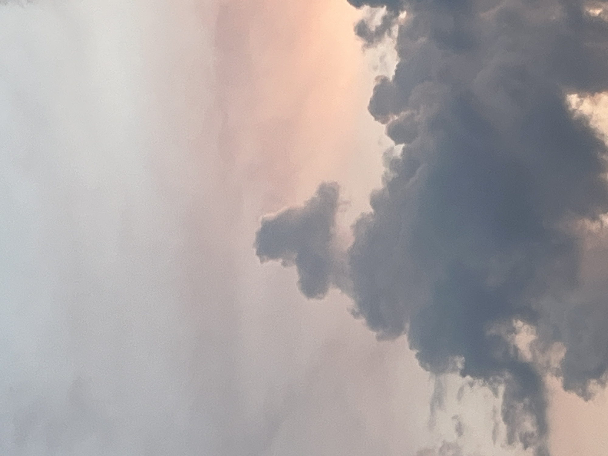The image displays a close-up view of a sky with clouds at dusk. The clouds are predominantly dark gray, with some lighter gray and pinkish hues visible towards the top right, indicating the setting sun. A peculiar formation of clouds in the center appears to resemble an animal, possibly a dog or a rabbit, in profile, facing left. The background sky transitions from a pale gray at the top to soft pink and orange tones near the horizon, which is not visible in the frame. There are no people, buildings, or any other identifiable objects or structures in the image, besides the clouds. The scene is entirely focused on the atmospheric conditions and the cloud formations. This image was taken in Garmisch-Partenkirchen, Germany, a location known for its mountainous terrain and clear skies, often resulting in dramatic cloud formations, especially during sunrise and sunset.