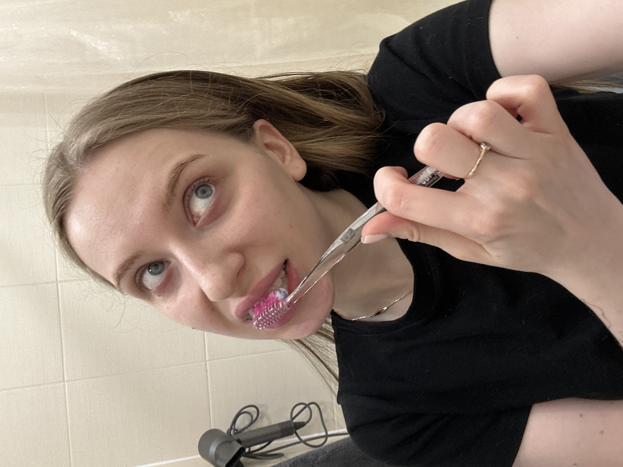 This image depicts a female individual brushing her teeth in an indoor bathroom environment. The subject, viewed from the chest up, has light brown hair and blue eyes, and is wearing a black short-sleeved top and a thin gold-colored necklace. She wears a gold-colored ring with a clear stone on her right ring finger. Her right hand holds a clear-handled toothbrush with pink bristles, positioned against her upper teeth. Her mouth is slightly open, and her gaze is directed upwards and to the viewer's left. The background features a tiled wall in a light beige color, with a black hairdryer hanging on a fixture and a dark gray towel partially visible below it. The precise location within the city of Chaikovsky, Russia, cannot be determined from the visual evidence.