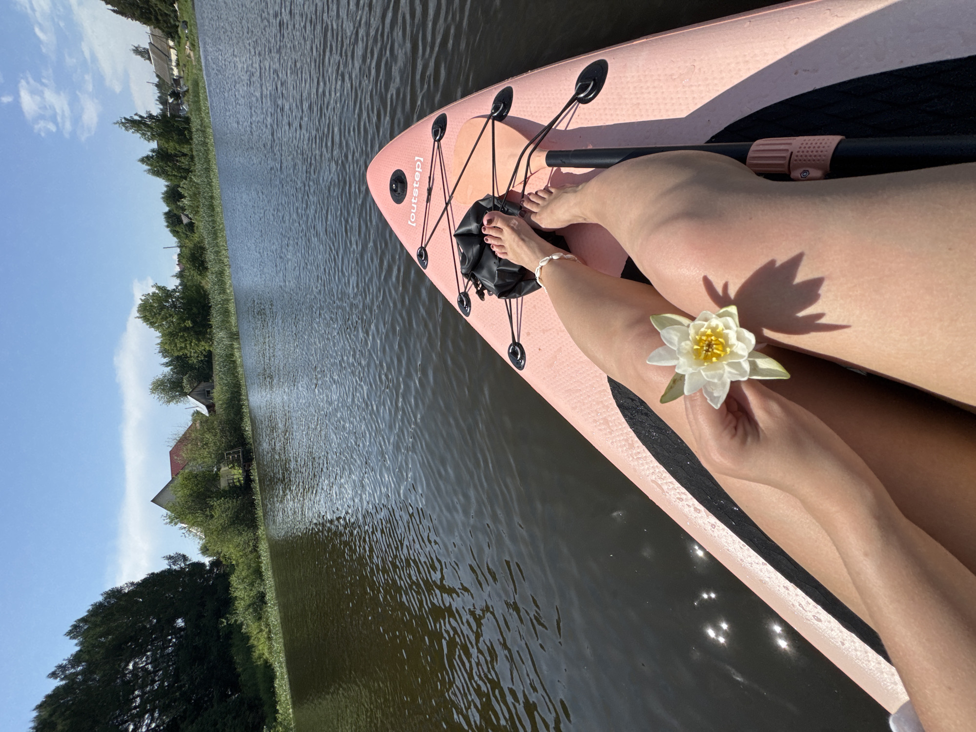 This media file depicts a person on a pink paddleboard on a calm body of water. The scene is set on a sunny day, likely in the afternoon, with a clear blue sky and scattered white clouds visible. The paddleboard is equipped with black bungee cords for storage and a black paddle handle is visible.

The person's legs are visible, one resting on the paddleboard and the other bent. They are wearing a seashell anklet on their right ankle. The person is holding a delicate white water lily in their right hand, with its petals slightly unfurled. A shadow of the water lily is cast onto their skin.

The background shows a lush green shoreline with trees and what appear to be houses, suggesting a rural or semi-rural location. The water is dark and reflects the sunlight, creating shimmering patterns on its surface. The overall mood is peaceful and serene, evoking a sense of relaxation and connection with nature.

The visible text on the paddleboard reads "[outstep]". The location is identified as Chastyye, Russia.