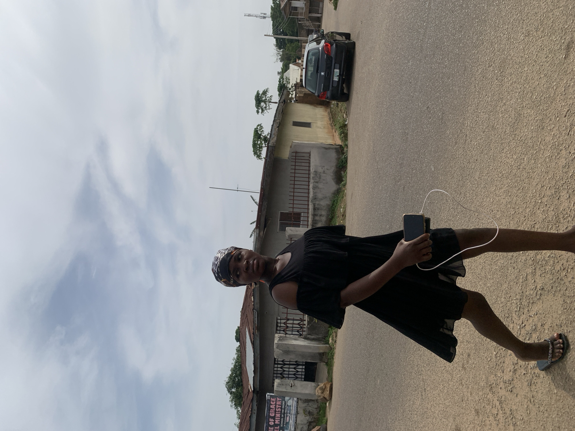 A young Black woman is walking on a dusty road in Jos, Nigeria. She is wearing a black off-the-shoulder dress and a headscarf with a black and white geometric pattern. She is holding a smartphone in her right hand, and a white wire, possibly for headphones or charging, is attached to it and drapes down. Her left foot is bare, while her right foot is wearing a dark sandal with embellishments. The road is paved but appears worn, with a sandy shoulder. To the left of the woman, a building with a sign that reads "THE OF GRACE MINISTRIES" is visible. The building has a corrugated metal roof and barred windows. Beyond this building, more structures and vegetation can be seen. On the right side of the road, a black Peugeot 308 car is parked. The sky is partly cloudy.