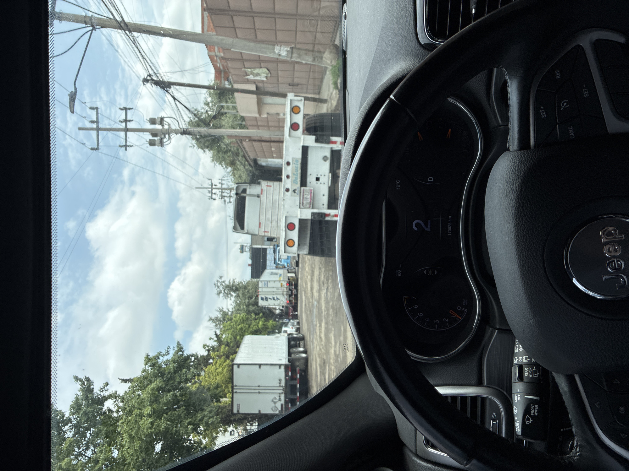 Captured from inside a vehicle, the photograph looks out onto a bustling street presumably within Cuautitlán Izcalli, Mexico. The perspective is from the driver's seat, with the vehicle's steering wheel prominently visible. A long line of trucks and trailers stretches ahead, implying a traffic jam or congested road. Power lines and buildings are evident in the background, adding to the urban setting. The dashboard indicates a speedometer reading of "2" and a temperature of 19°C. The brand "Jeep" is prominently displayed on the steering wheel.