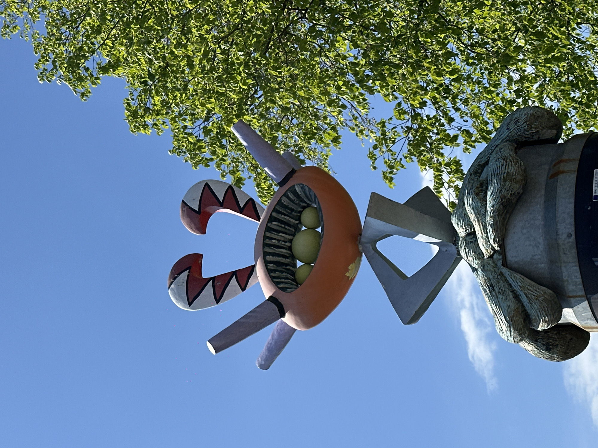 The image depicts a colorful, abstract sculpture against a backdrop of a bright blue sky and green tree foliage. The sculpture consists of an orange, circular body with white and red zigzag patterns on its appendages, and lavender-colored protrusions. Inside the circular body, there are several round, yellowish objects. The sculpture is mounted on a metallic pole that has a bronze-colored, organic-looking design wrapped around it. The photo was taken from a low angle, looking up towards the sculpture and the sky. The location is in Amsterdam, Netherlands.
