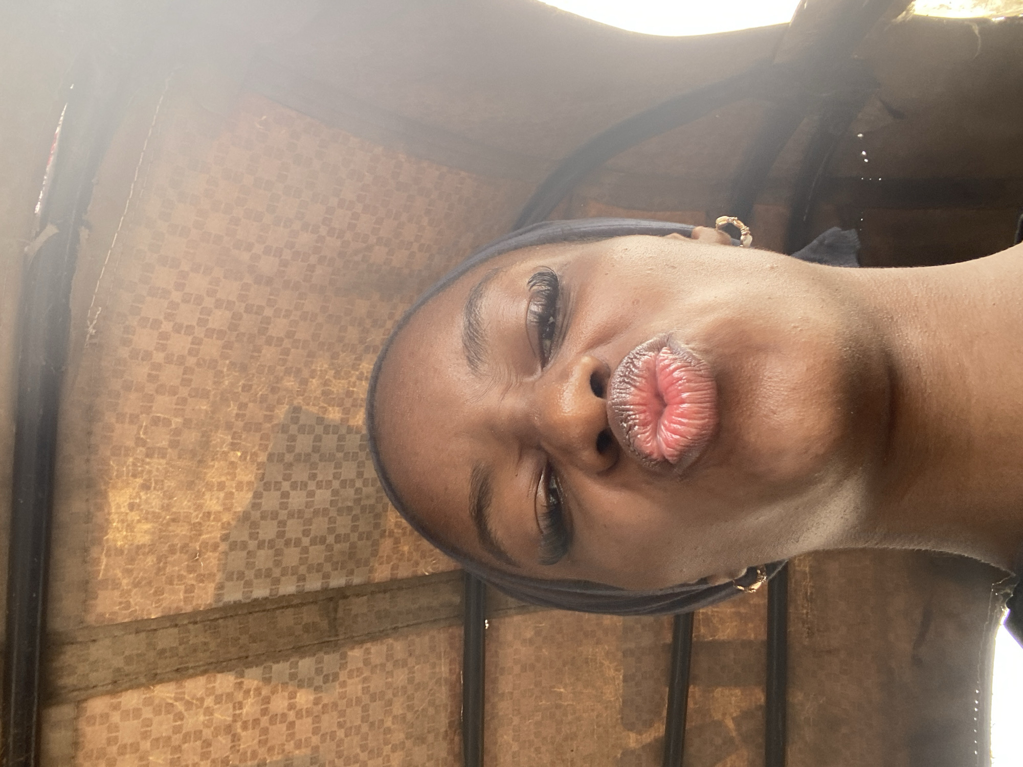 A close-up, low-angle, headshot of a young woman in a vehicle. She is looking directly at the camera with her lips pursed as if blowing a kiss. The woman has dark skin, dark eyes, and her eyebrows are well-defined. She is wearing a dark headscarf and a gold hoop earring in her right ear. Her expression appears playful and affectionate.
The background is the interior of what appears to be a small vehicle, possibly a tuk-tuk, given the patterned upholstery and the visible metal bars. The upholstery is a light brown or tan color with a repeating diamond pattern. The lighting suggests it is daytime, with sunlight filtering in, creating a bright, somewhat hazy effect and casting shadows. The scene is set indoors, within the confines of the vehicle. There is no visible text in the image. The overall mood is casual and intimate. The location context of Jos, Nigeria, suggests an urban or semi-urban environment.