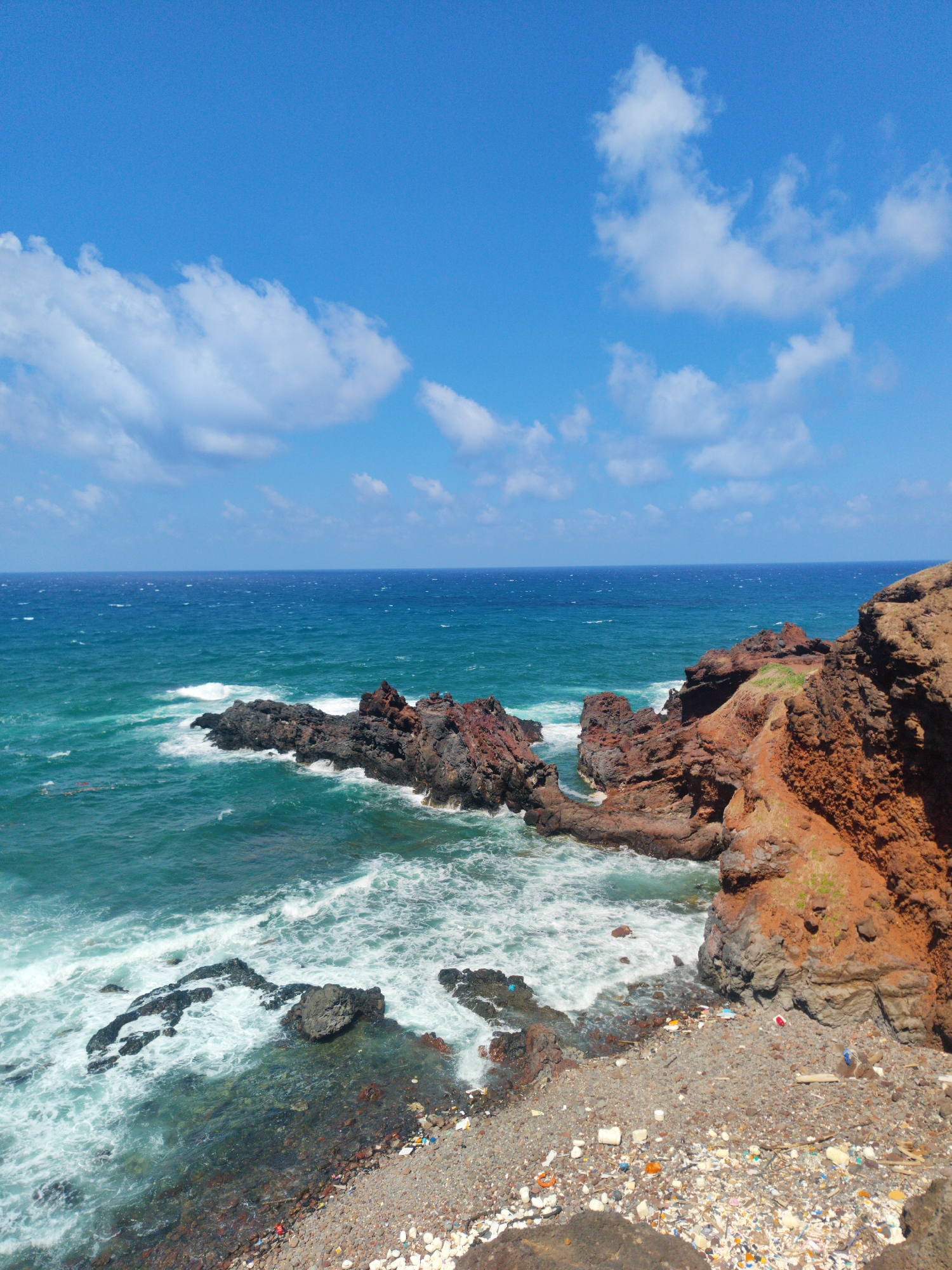 The image depicts a rugged coastal scene in Phú Quý Special Administrative Region, Vietnam. The turquoise ocean is agitated with white-capped waves crashing against dark, jagged rock formations. A prominent headland of reddish-brown rock dominates the right side of the frame, contrasting with the darker, more weathered rocks in the water. The rocky beach is littered with debris and trash, indicating potential environmental pollution. The sky above is a bright blue, dotted with scattered white clouds. The overall impression is of a naturally beautiful location marred by human impact.