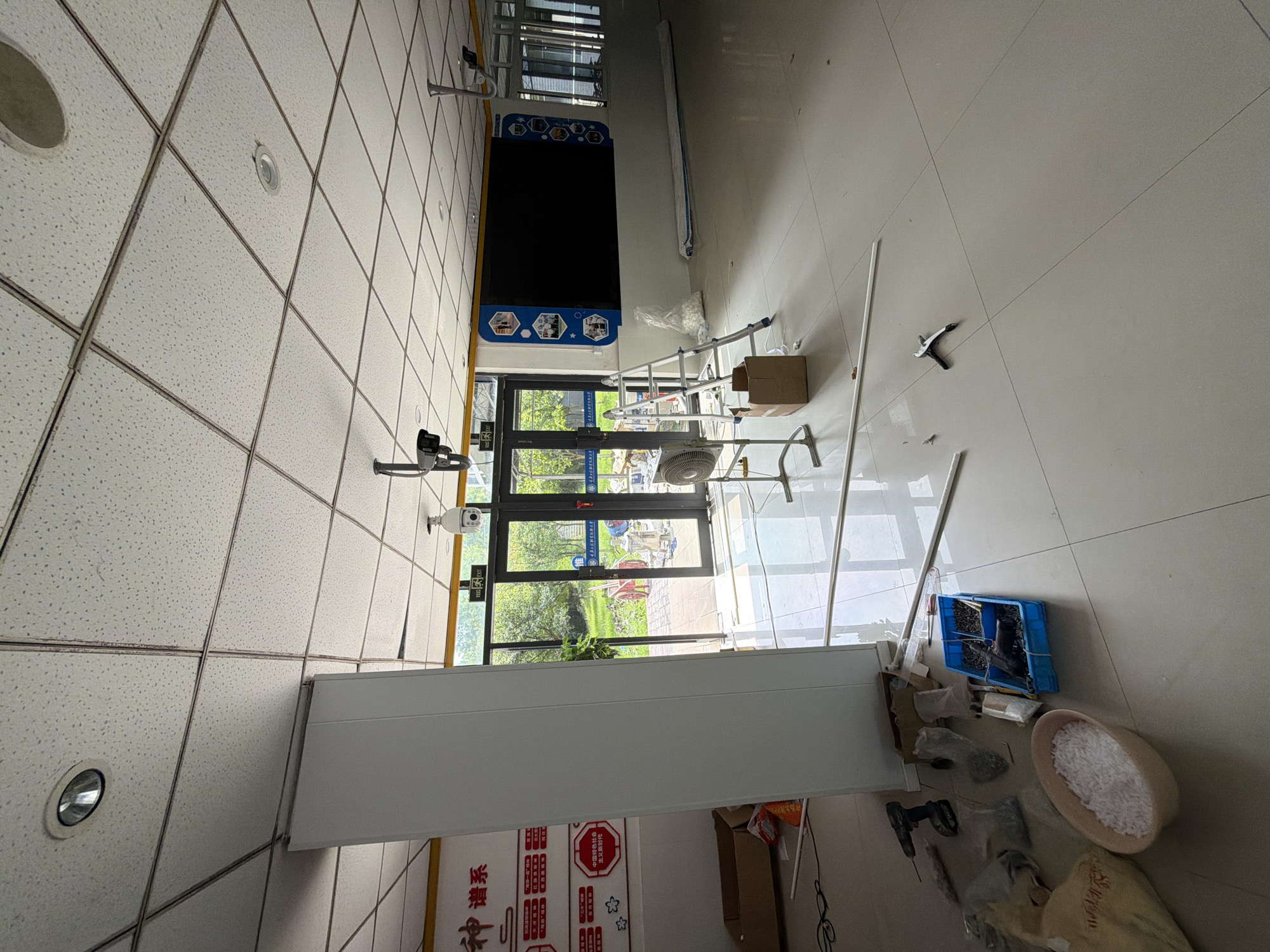 The image depicts an indoor space that appears to be undergoing renovation or setup, possibly a classroom or meeting room, in the city of Jiangjin, China. The ceiling is a standard drop ceiling with fluorescent lighting fixtures. White, rectangular panels are visible on the left side, suggesting a newly installed partition or wall.
In the foreground, various construction materials and tools are scattered on the tiled floor. These include a cordless drill, a blue plastic container filled with screws and nails, a pair of pliers, a bowl of what appears to be small white fasteners or insulation, and a yellow plastic bag. A tall, white partition stands prominently on the left.
Towards the middle ground, a portable fan is positioned near a set of glass double doors that lead outside to a green, natural environment with trees. A ladder is set up near the doors, leaning against the wall where a large, dark digital display screen is mounted. The screen is adorned with decorative hexagonal shapes and images on its blue surround. Two security cameras are visible, one mounted on the ceiling near the entrance and another above the digital display. An "EXIT" sign with an illuminated green arrow is also present near the double doors. The overall impression is a scene of transition, with ongoing work in a relatively empty space.