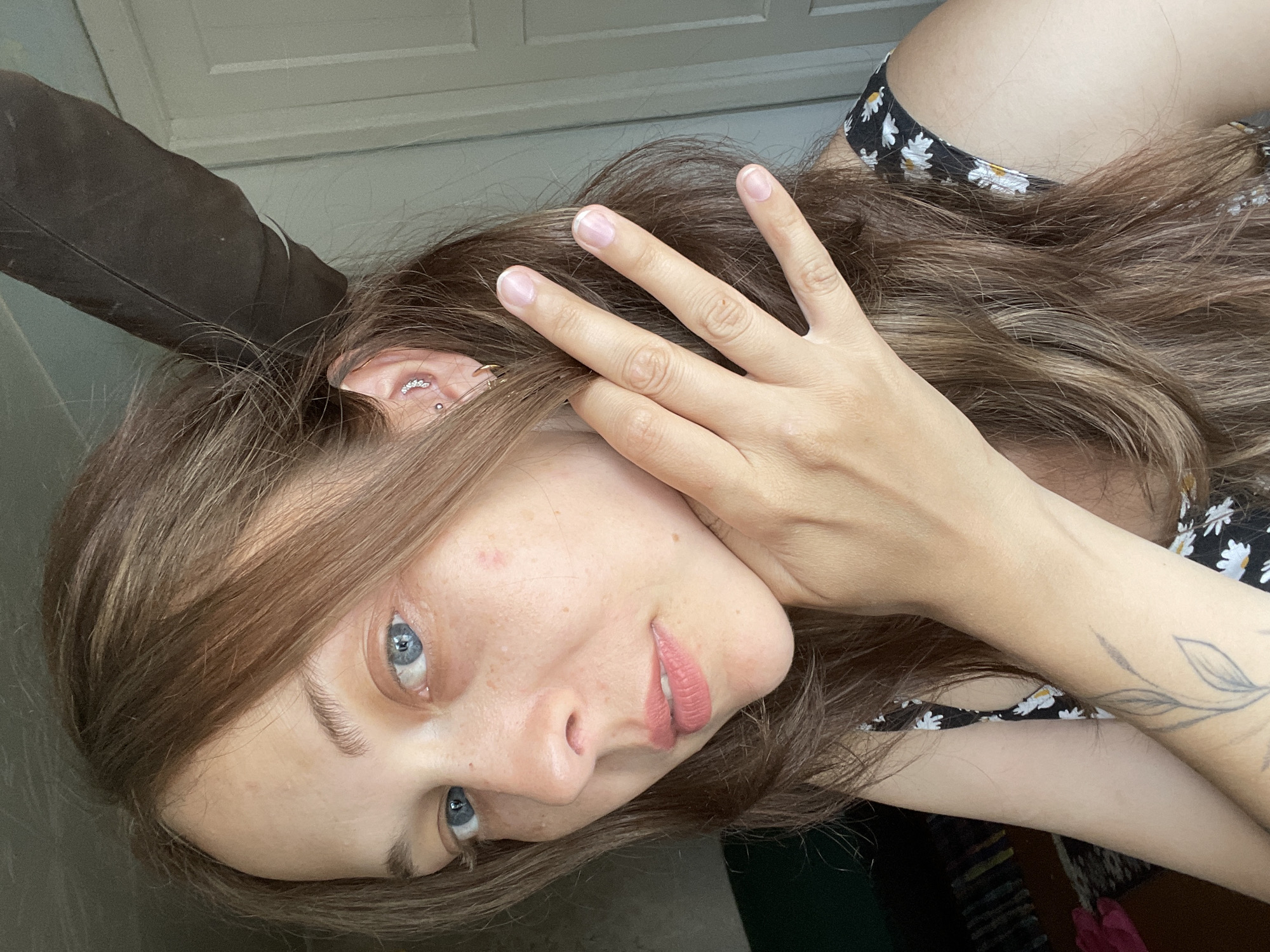 The image is a close-up, medium-shot selfie of a woman with light skin, brown hair, and blue eyes. She is looking towards the camera with her left hand touching her neck and her right hand near her face, with fingers splayed. She is wearing a black floral dress. There is a large, dark feather positioned behind her head and to the right. The background appears to be an indoor setting with a wall and a door visible on the right. The lighting suggests it is daytime. No specific location in a city is discernible.
