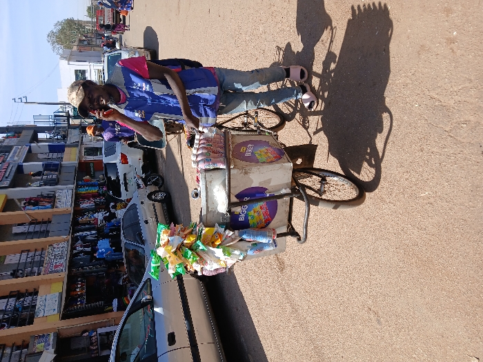The image depicts an urban street scene in Unknown, Unknown, under bright daylight. A male individual, identified as a street vendor, stands slightly right of center. He has dark skin, a beard, and wears a tan baseball cap, a pink t-shirt, a blue and white vest, light blue jeans, and pink open-toed sandals. He is holding a dark mobile phone to his right ear, engaged in a conversation. He stands beside a customized bicycle with a large, rectangular, light-colored cart attached to its front, supported by two wheels. This cart is adorned with numerous packaged snacks hanging from its left side and several pink-labeled bottled beverages on its top surface, displaying "BIGGER" and "BUDGET" branding. The street surface is reddish-brown asphalt or dirt, showing distinct shadows. In the background, multi-story commercial buildings line the left side of the street, featuring shops with clothing items displayed outside. Several parked vehicles, including a silver sedan, a gold sedan, a white SUV, and a motorcycle, are visible. Additional indistinct individuals are present in the distant background, walking along the street.
